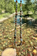 Current necklace by Auréad Designs held against the multicolored argillite riverbed of Glacier National Park, with Indian agate beads in full natural color variation reflecting the reds, greens, and tans of the stone below