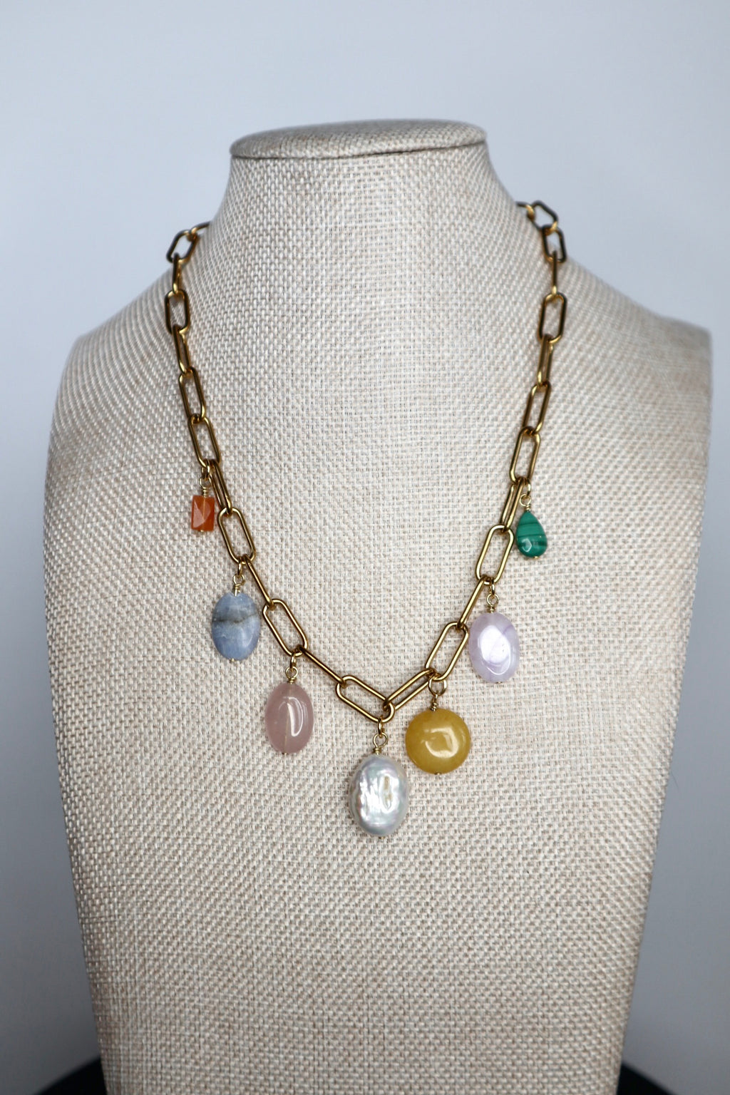 Elavene 18K gold-plated paperclip chain necklace by Auréád Designs displayed on a linen bust form, featuring seven mixed gemstone charm drops including orange aventurine, blue lace agate, rose quartz, freshwater pearl, yellow jade, lavender amethyst, and malachite