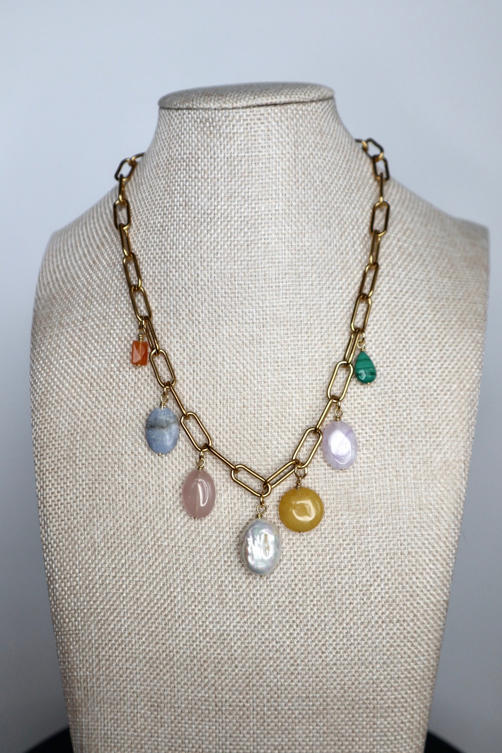 Elavene 18K gold-plated paperclip chain necklace by Auréád Designs displayed on a linen bust form, featuring seven mixed gemstone charm drops including orange aventurine, blue lace agate, rose quartz, freshwater pearl, yellow jade, lavender amethyst, and malachite