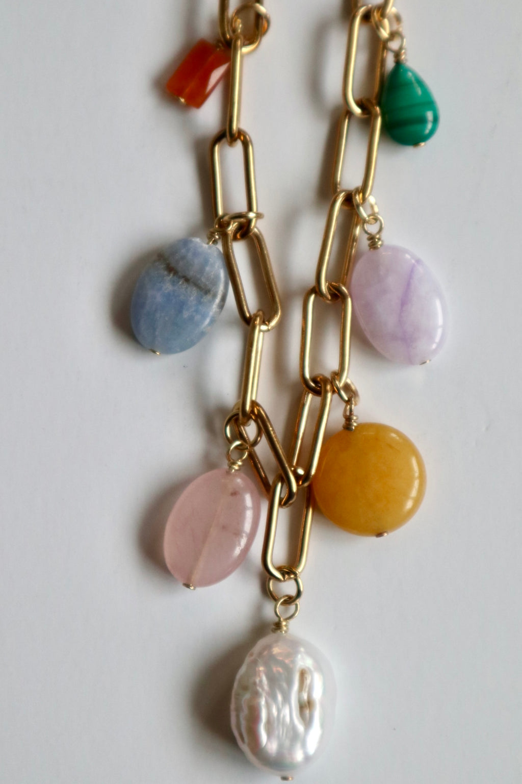 Close-up detail of the Elavene necklace by Auréád Designs laid flat on a white surface, showing seven mixed gemstone charm drops of orange aventurine, blue lace agate, rose quartz, freshwater pearl, yellow jade, lavender amethyst, and malachite hanging from the gold paperclip chain