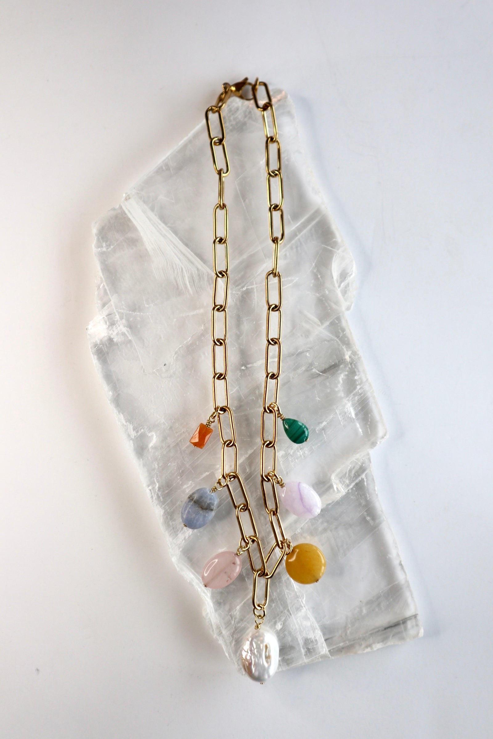 Elavene 18K gold-plated paperclip chain necklace by Auréád Designs laid flat on a white selenite slab, displaying seven mixed gemstone charm drops including orange aventurine, blue lace agate, rose quartz, freshwater pearl, yellow jade, lavender amethyst, and malachite