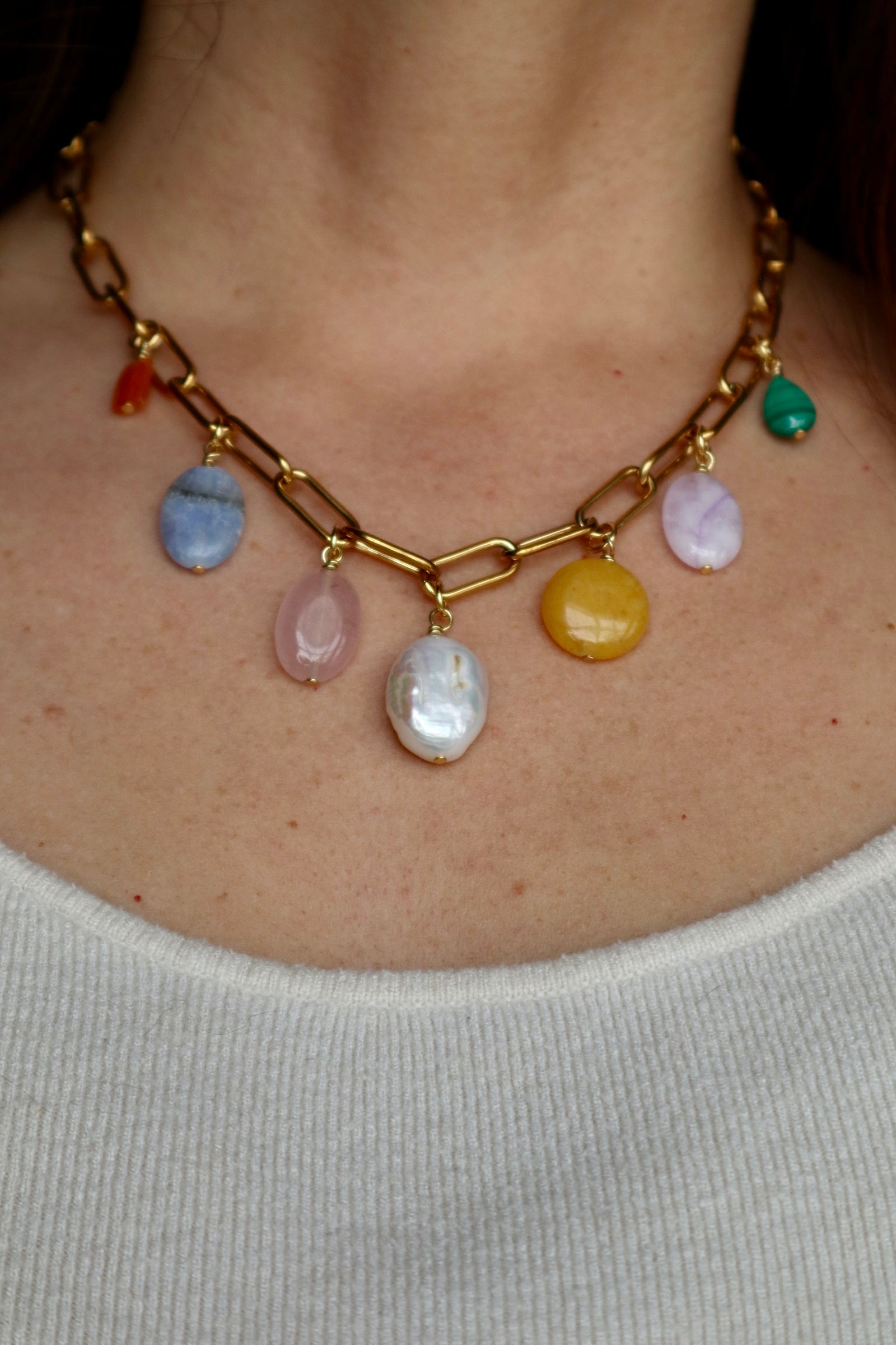 Elavene 18K gold-plated paperclip chain necklace by Auréád Designs worn on a bare neckline over a white top, displaying seven mixed gemstone charm drops including orange aventurine, blue lace agate, rose quartz, freshwater pearl, yellow jade, lavender amethyst, and malachite