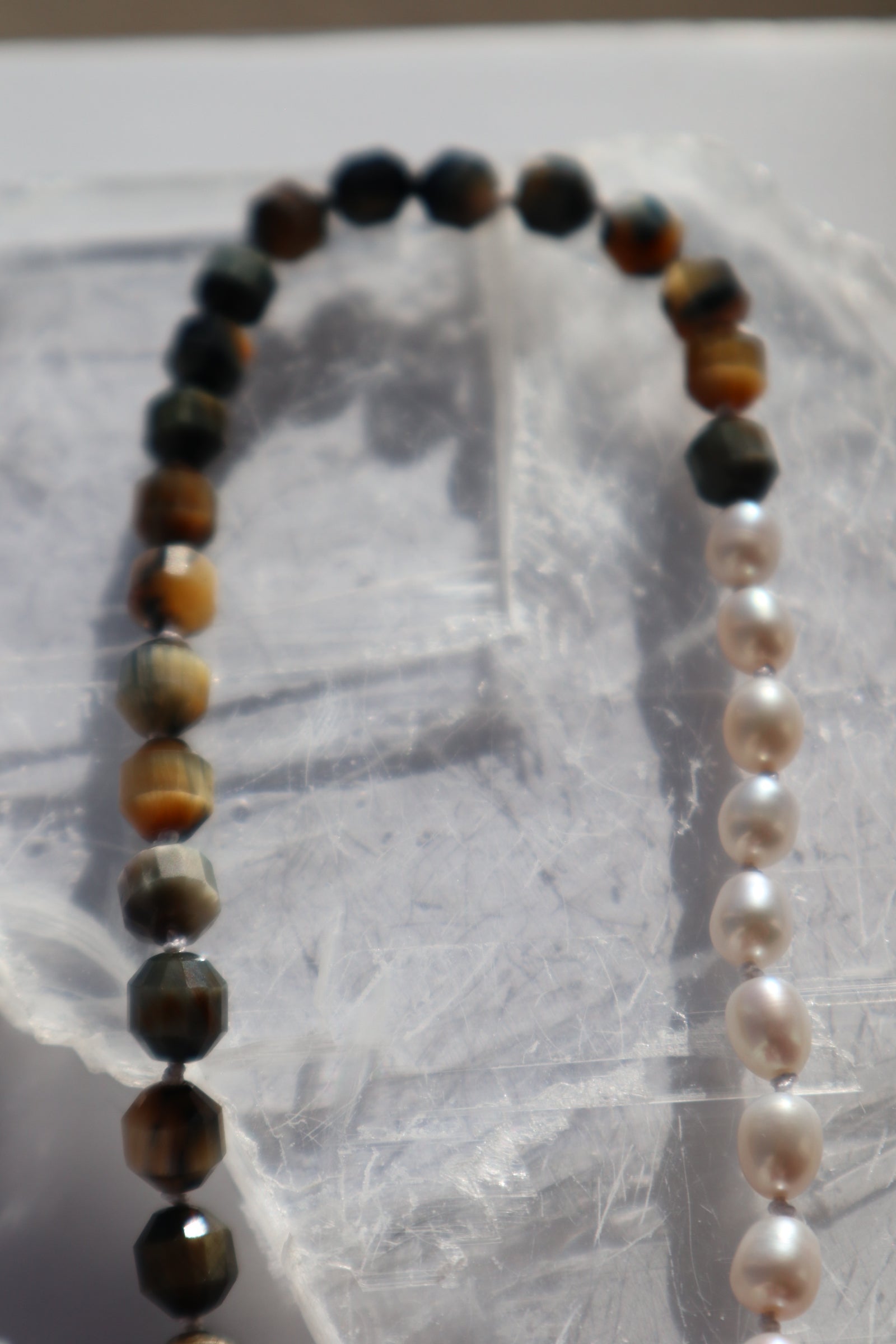 Close-up of the Equina necklace on a white selenite slab, showing the asymmetrical arrangement with deep blue tiger eye tube beads on one side and luminous oval freshwater pearls on the other