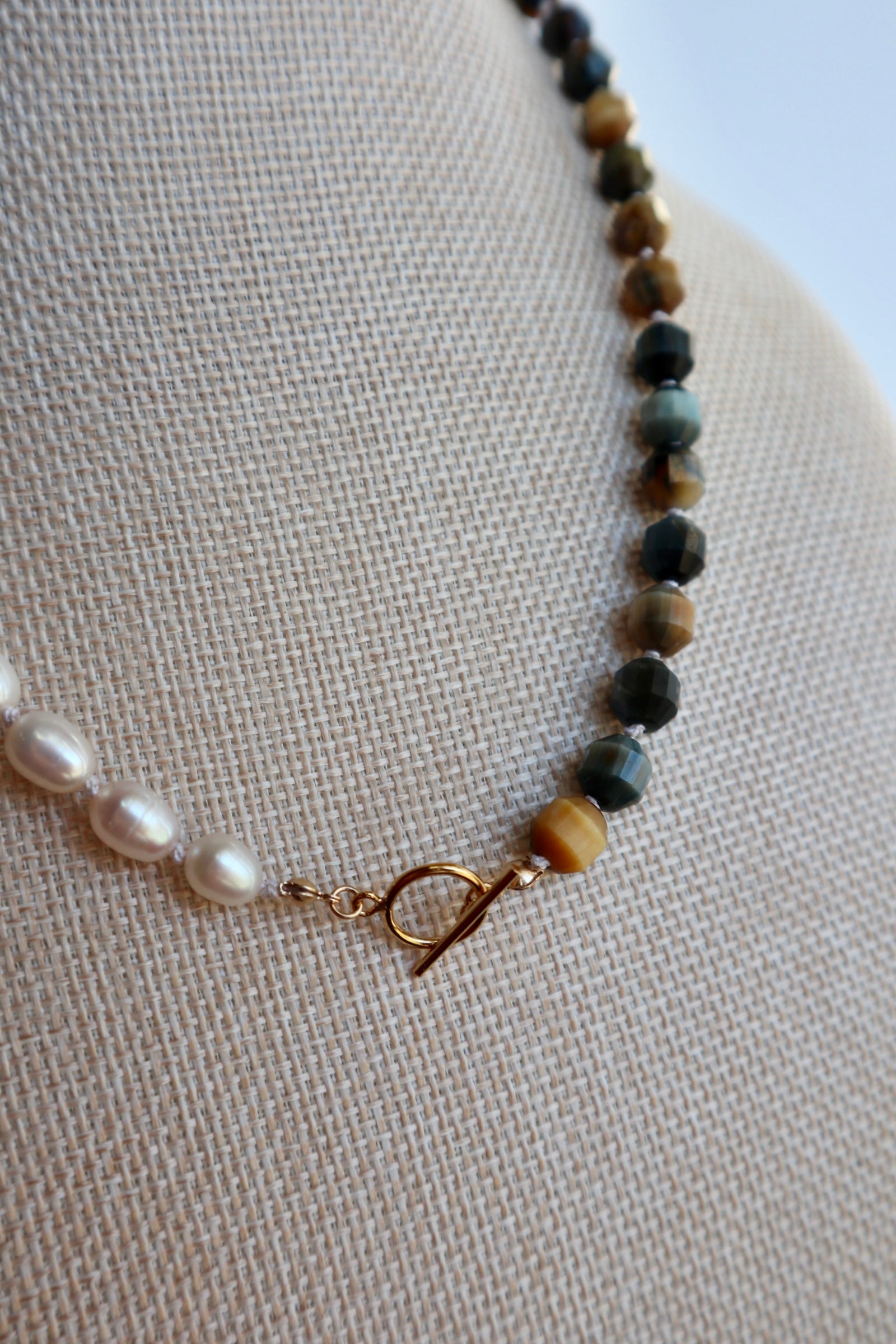 Equina necklace by Auréad Designs displayed on a linen bust form, highlighting the asymmetrical arrangement of deep blue tiger eye beads and oval freshwater pearls with the 14k gold-filled toggle clasp