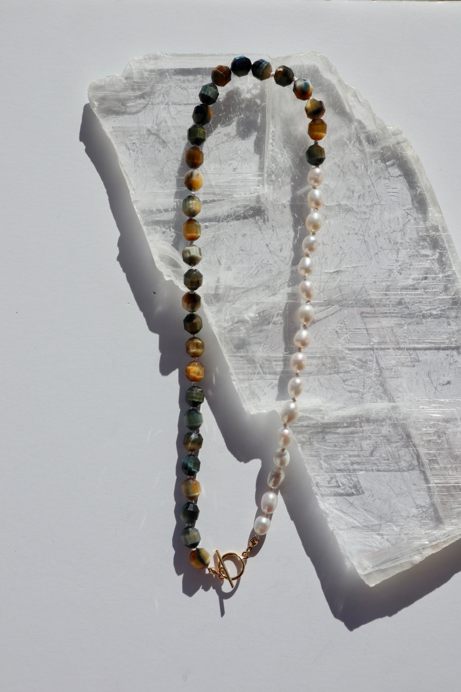 Equina necklace by Auréad Designs, featuring blue tiger eye tube beads and oval freshwater pearls on a white silk-knotted strand with a 14k gold-filled toggle clasp, laid flat on a white selenite slab