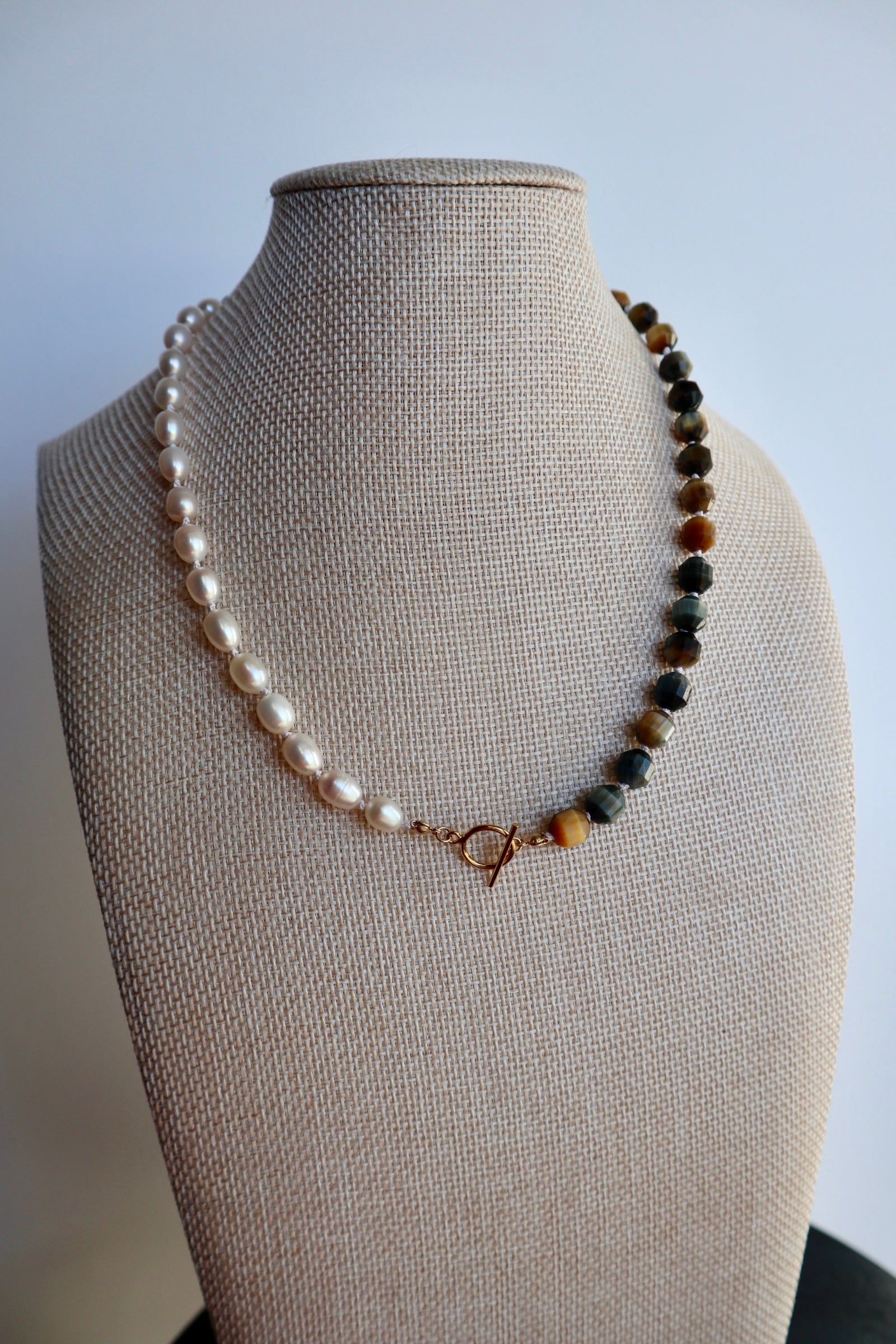 Equina necklace by Auréad Designs on linen bust, showing the asymmetrical arrangement of blue tiger eye tube beads and freshwater pearls on a white silk-knotted strand
