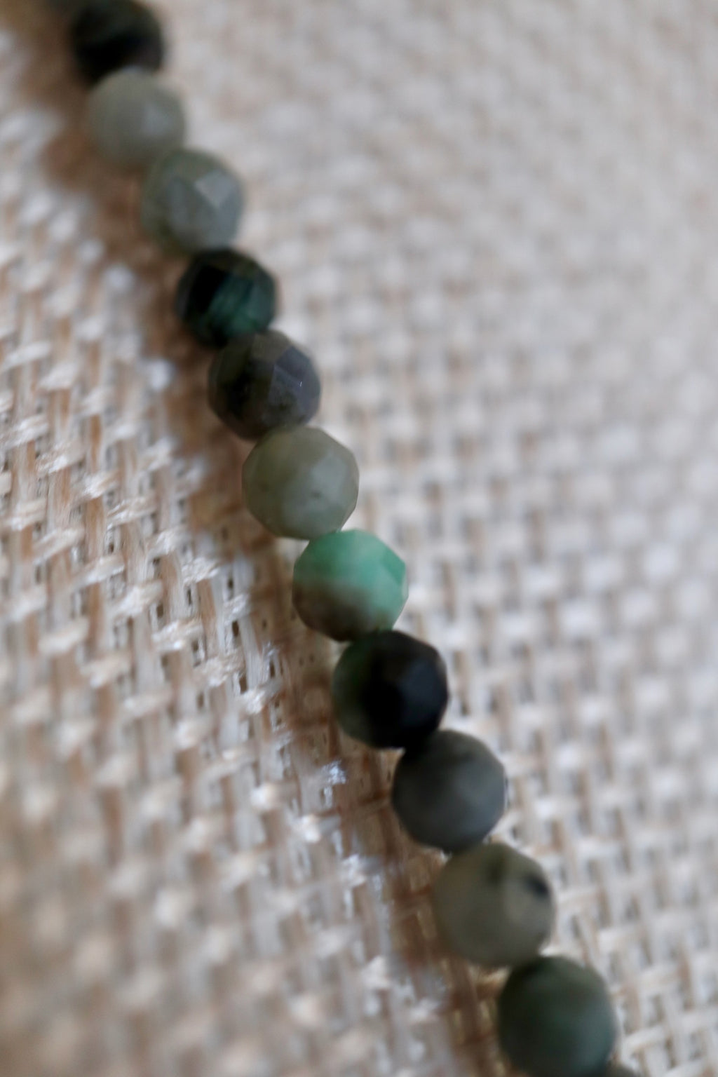 Auréad Designs Esmira necklace close-up of faceted emerald beads on linen bust form, showing natural variation in green tones from pale sage to deep forest