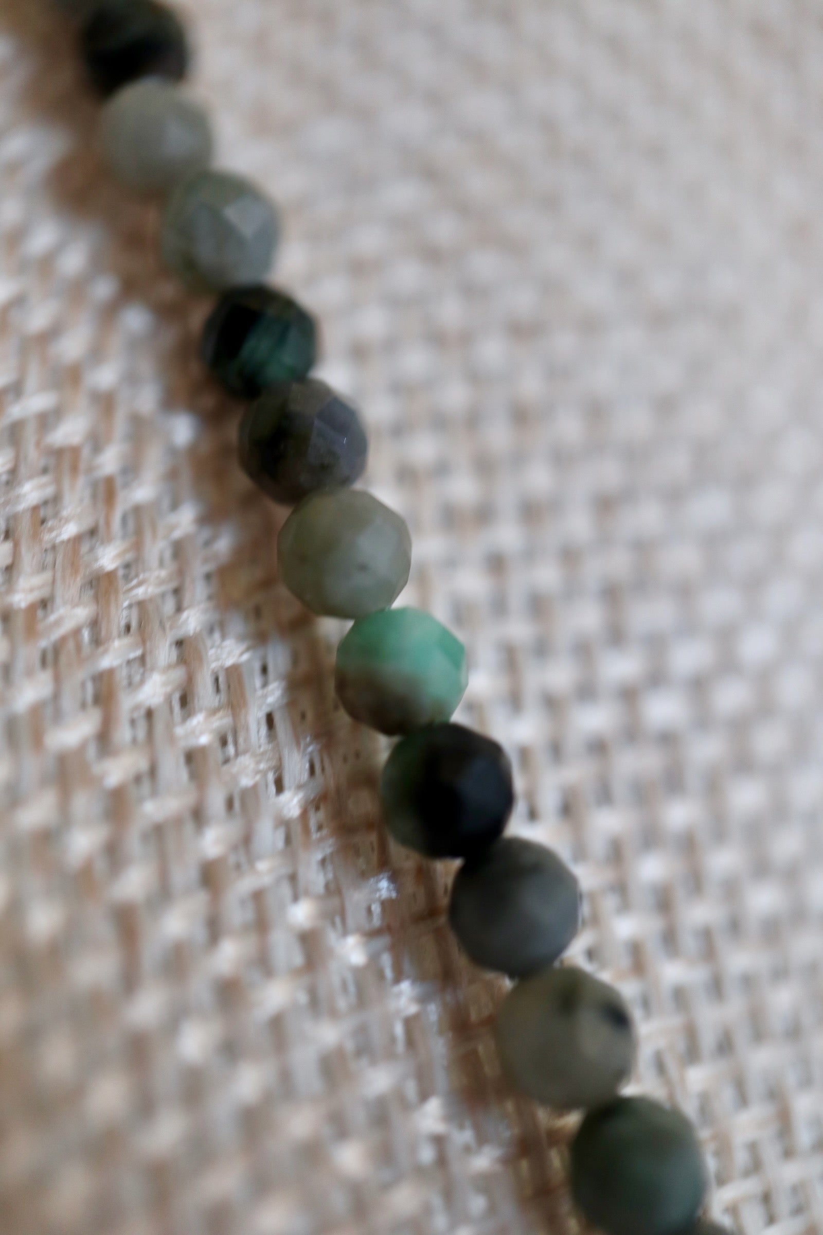 Auréad Designs Esmira necklace close-up of faceted emerald beads on linen bust form, showing natural variation in green tones from pale sage to deep forest