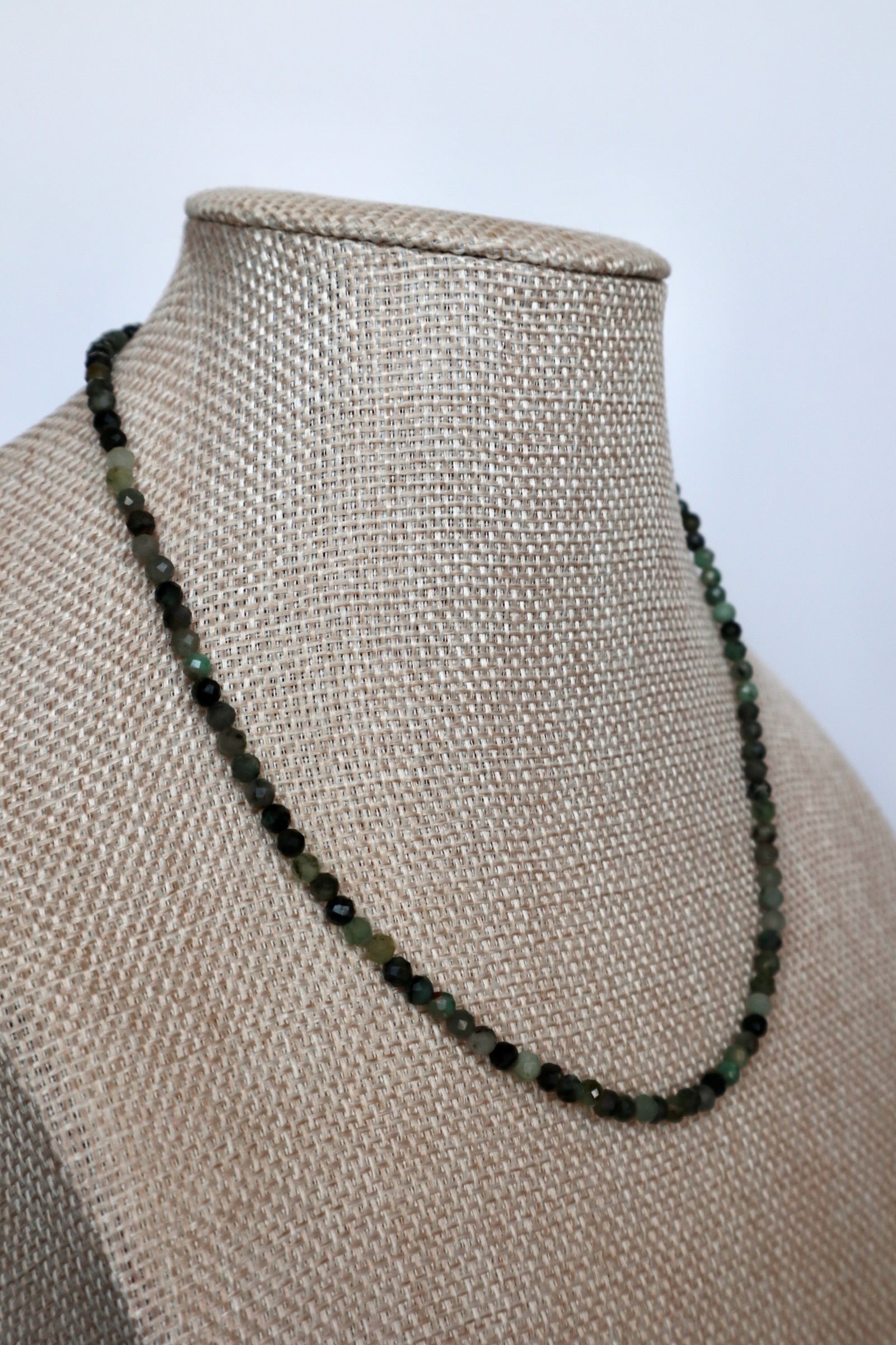 Auréad Designs Esmira necklace draped at an angle on linen bust form, showing full strand of 3mm faceted emerald beads against neutral linen texture