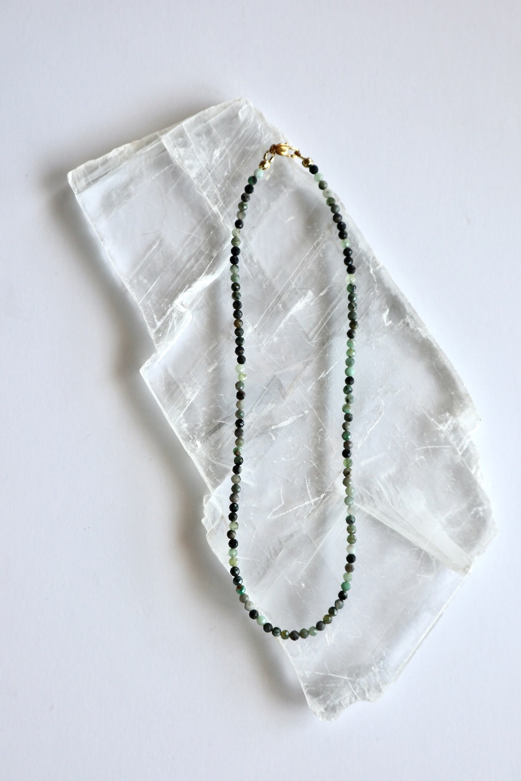 Auréad Designs Esmira necklace laid flat on white selenite slab, showing full strand of 3mm faceted round emerald beads with gold stainless steel lobster clasp