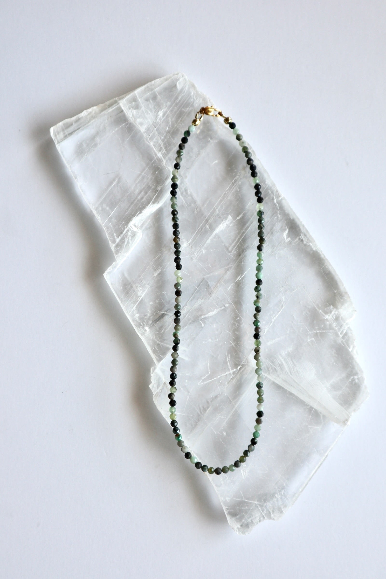 Auréad Designs Esmira necklace laid flat on white selenite slab, showing full strand of 3mm faceted round emerald beads with gold stainless steel lobster clasp
