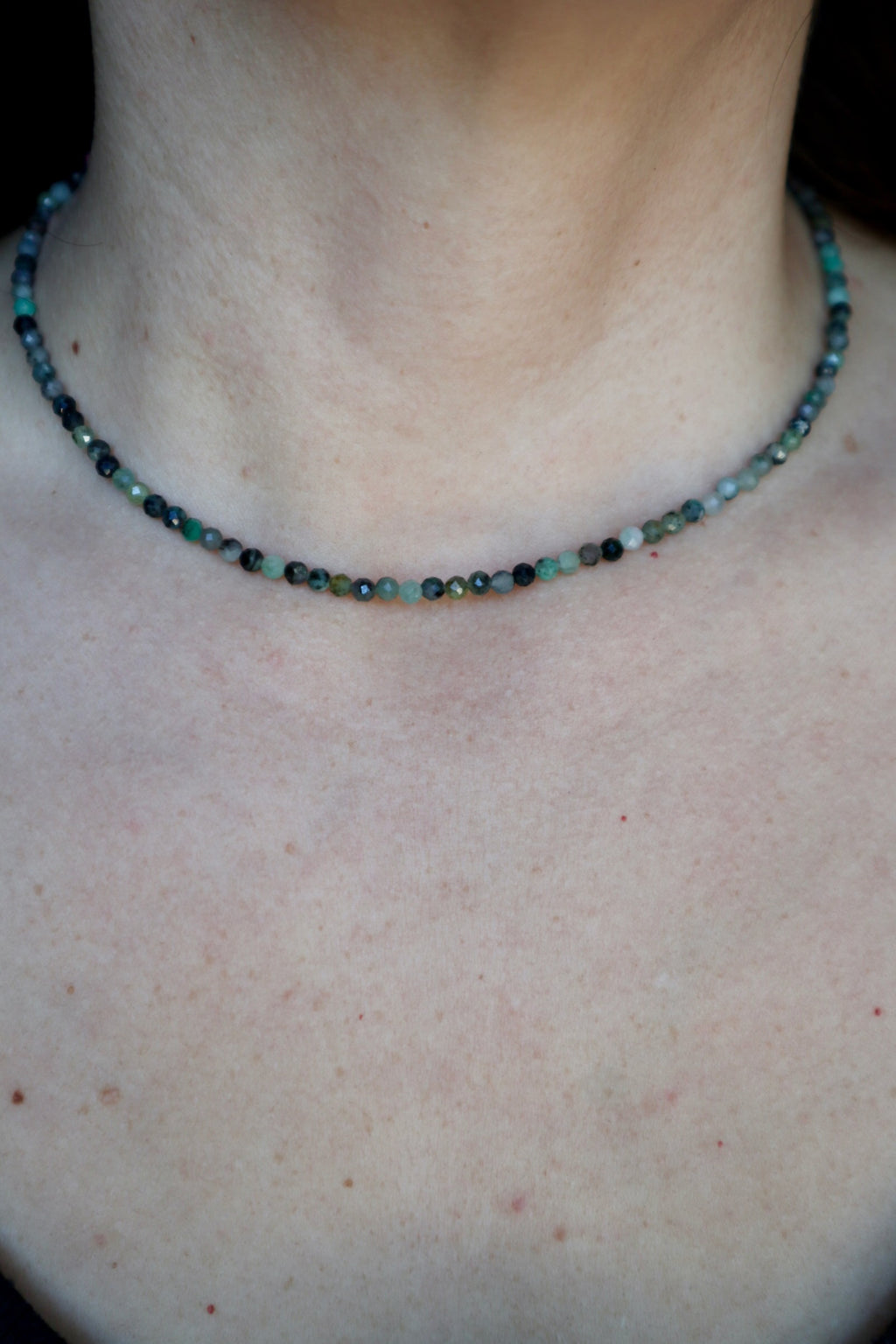 Auréad Designs Esmira necklace worn at collarbone length, showing faceted emerald bead strand against bare skin with natural color variation from deep forest green to pale sage visible across the strand
