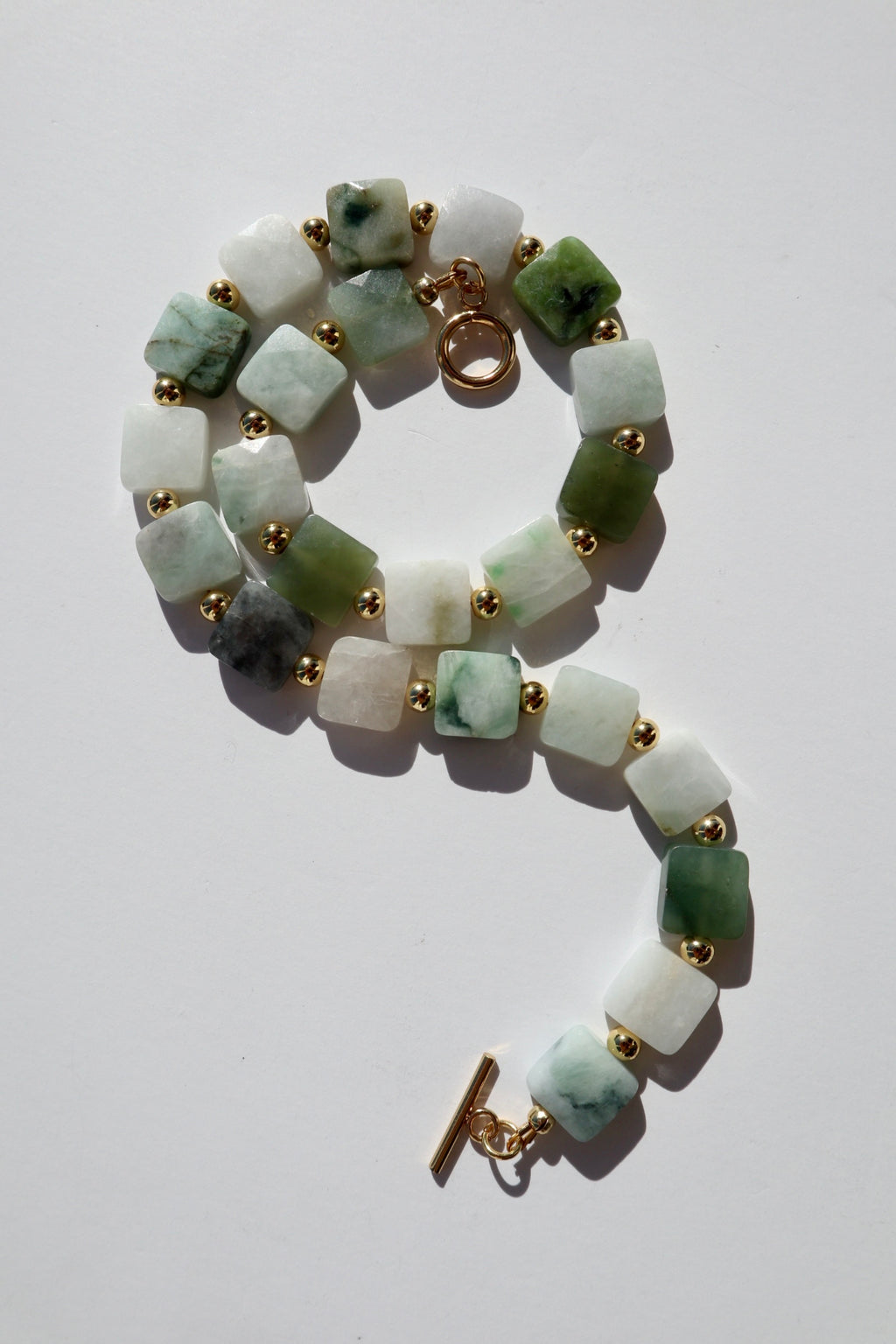 Everra necklace by Auréad Designs coiled on a white background, showing the full strand of square nephrite jade beads with natural color variation from deep green to pale celadon and white, with gold stainless steel spacers and toggle clasp