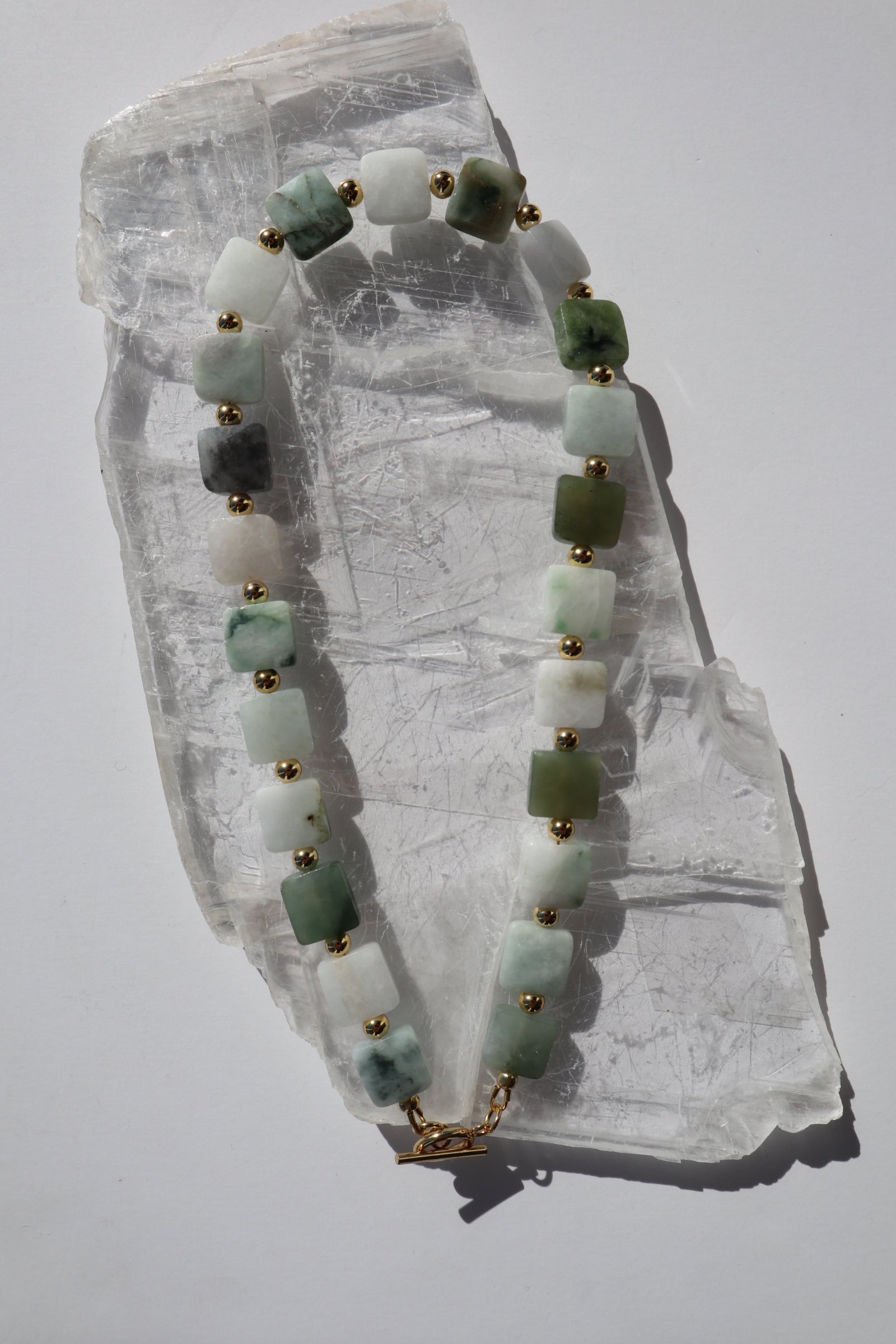 Everra necklace by Auréad Designs, featuring square nephrite jade gemstone beads and gold stainless steel spacer beads with a gold toggle clasp, laid flat on a white selenite slab