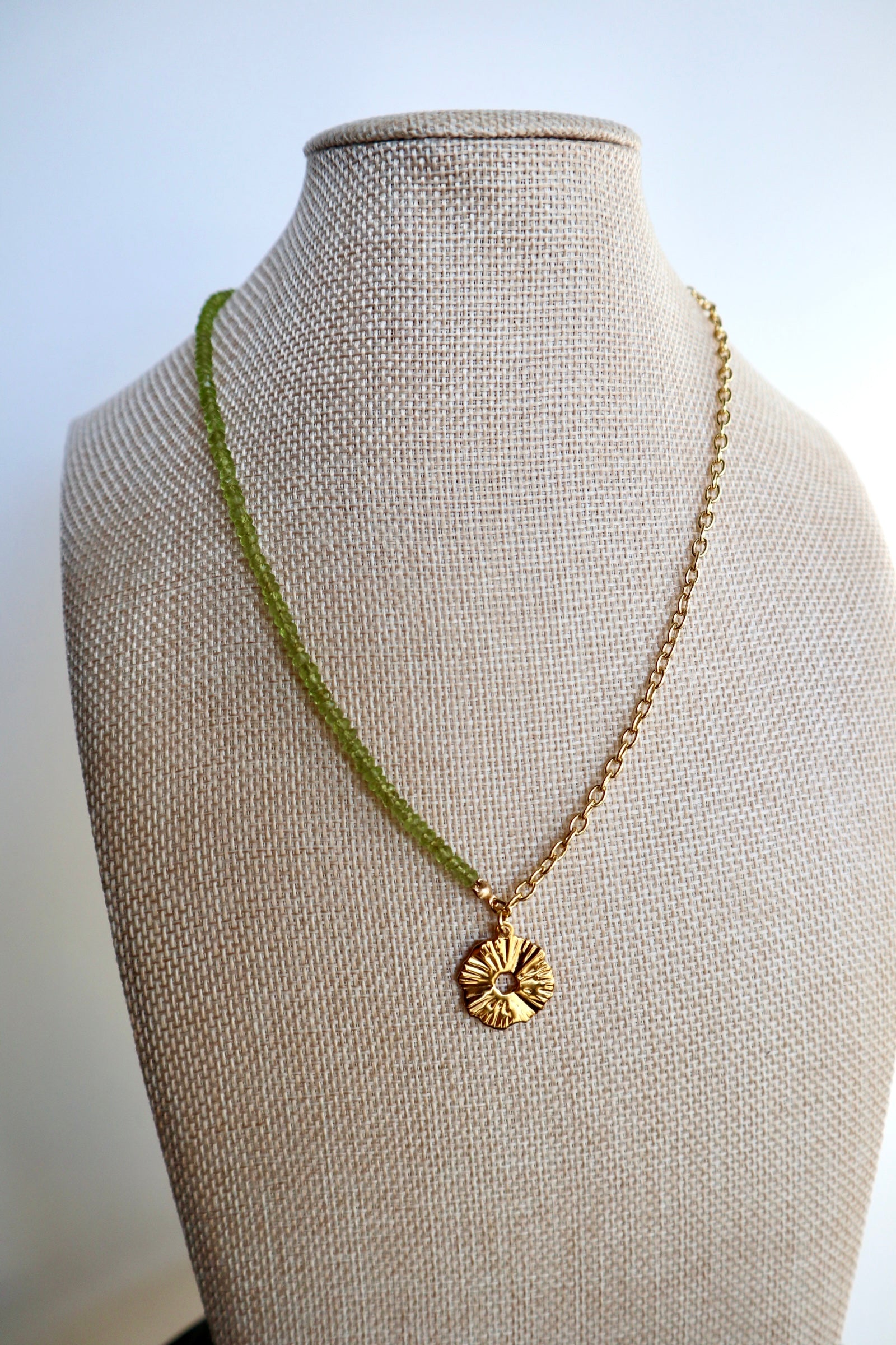 Florae necklace by Auréad Designs displayed on a linen bust form, showing the full asymmetrical design with faceted peridot beads on one side and a gold-filled cable chain on the other, with the flower pendant at the center