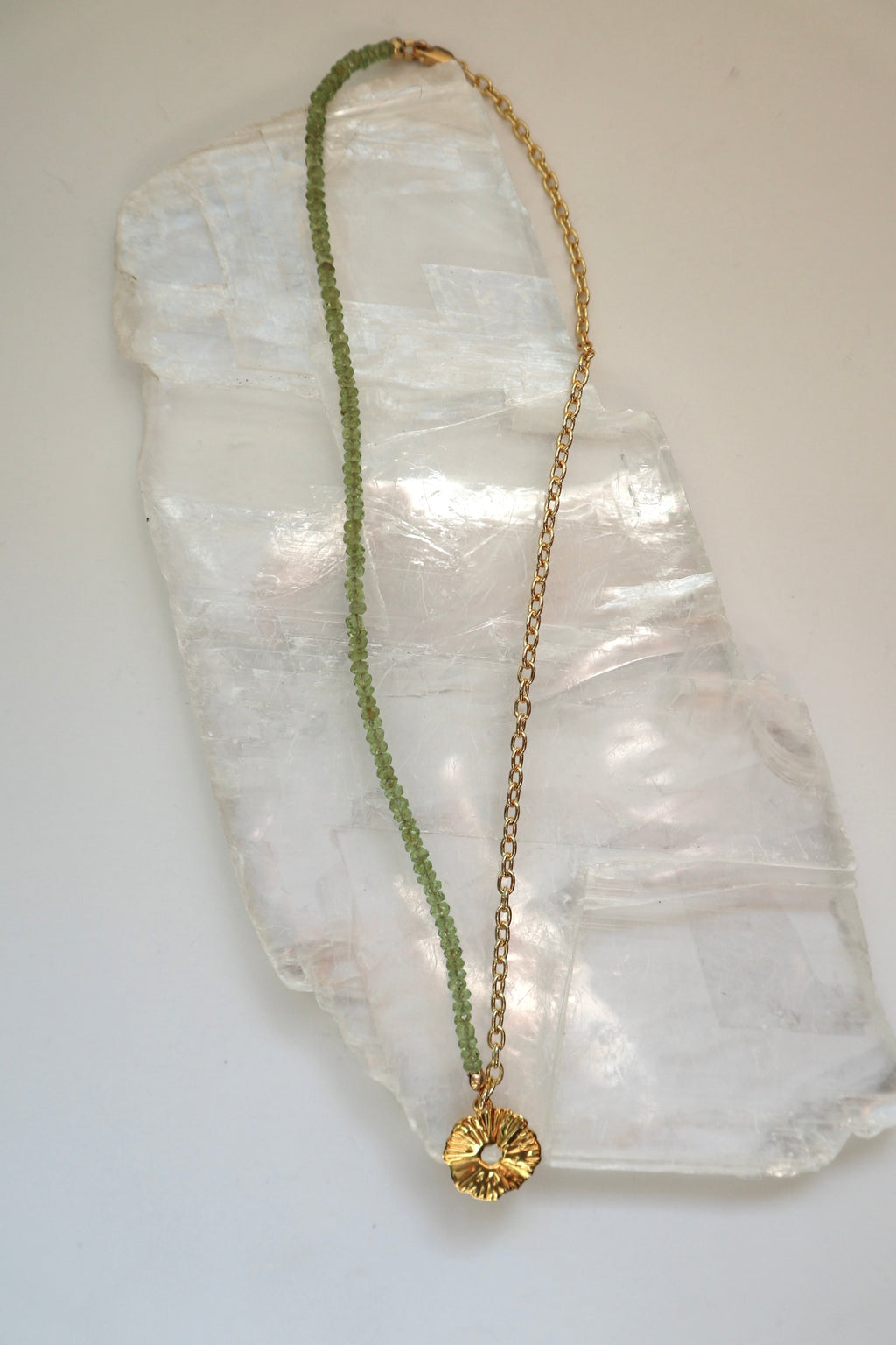 Florae necklace by Auréad Designs, featuring faceted peridot gemstone beads, a 14k gold-filled cable chain, and a gold flower pendant with a lobster clasp, laid flat on a white selenite slab