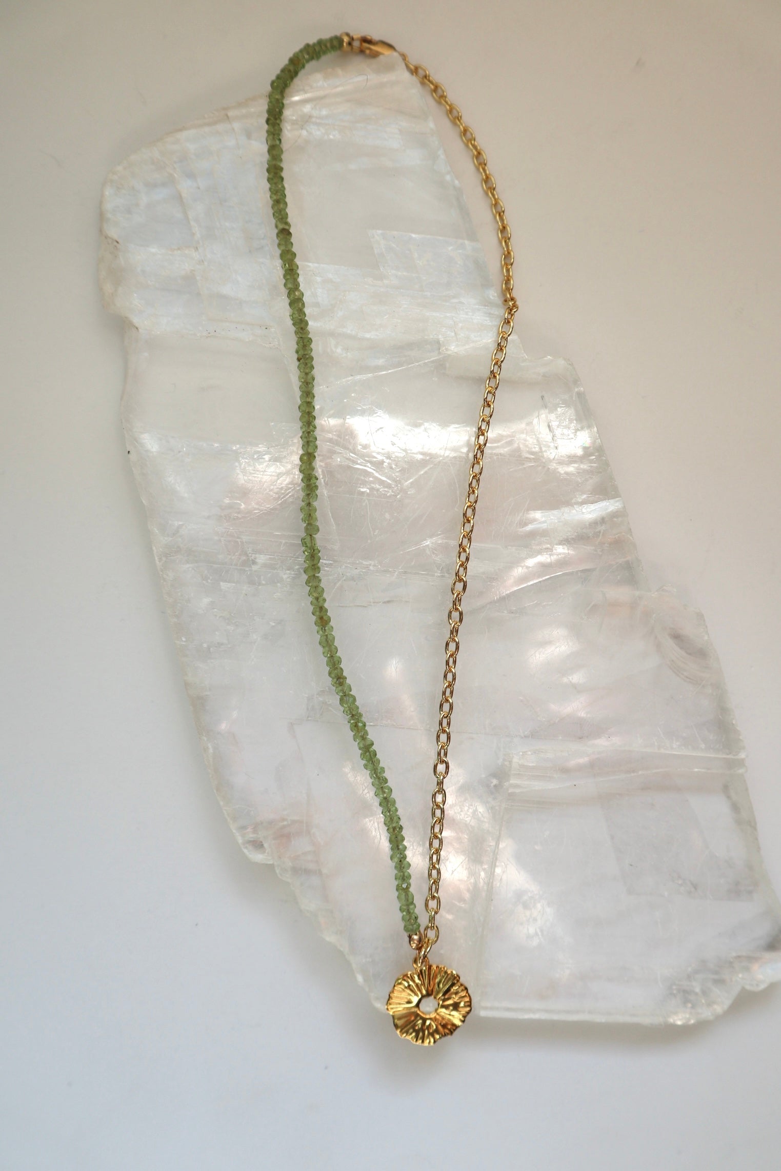 Florae necklace by Auréad Designs, featuring faceted peridot gemstone beads, a 14k gold-filled cable chain, and a gold flower pendant with a lobster clasp, laid flat on a white selenite slab