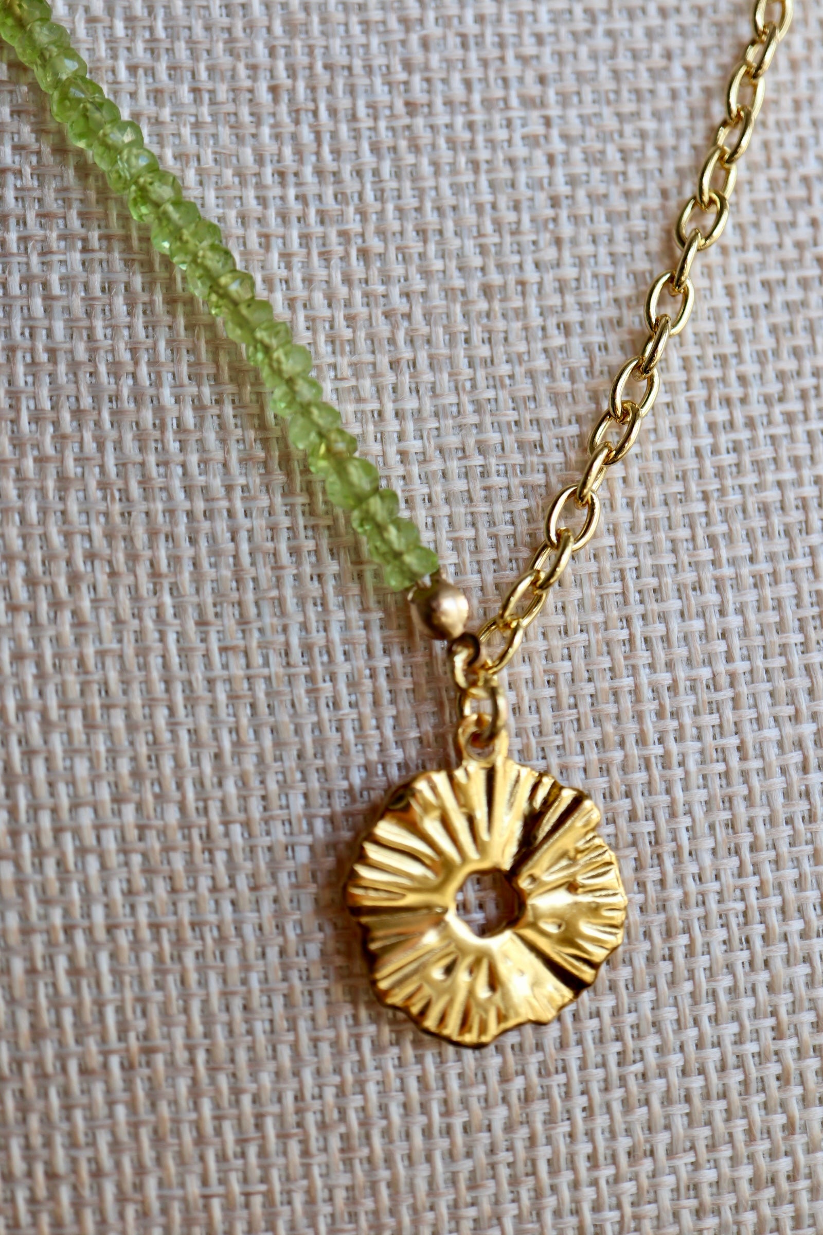 Close-up of the Florae necklace on a linen bust form, showing the ribbed sculptural PVD gold flower pendant at the junction of the faceted peridot bead strand and gold-filled cable chain