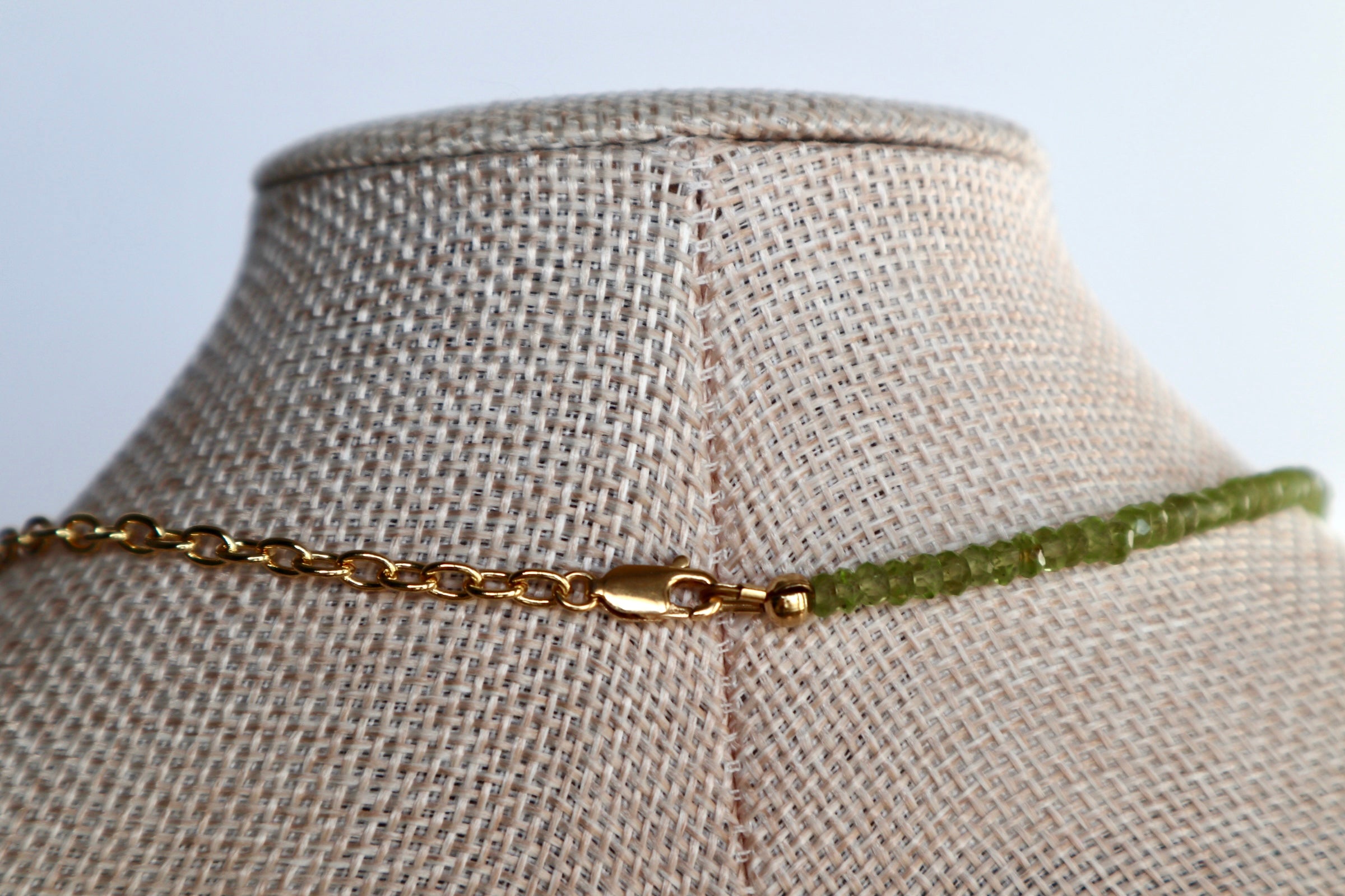 Close-up of the Florae necklace on a linen bust form showing the back of the piece, with the 14k gold-filled lobster clasp and the contrast between the gold cable chain and faceted peridot bead strand