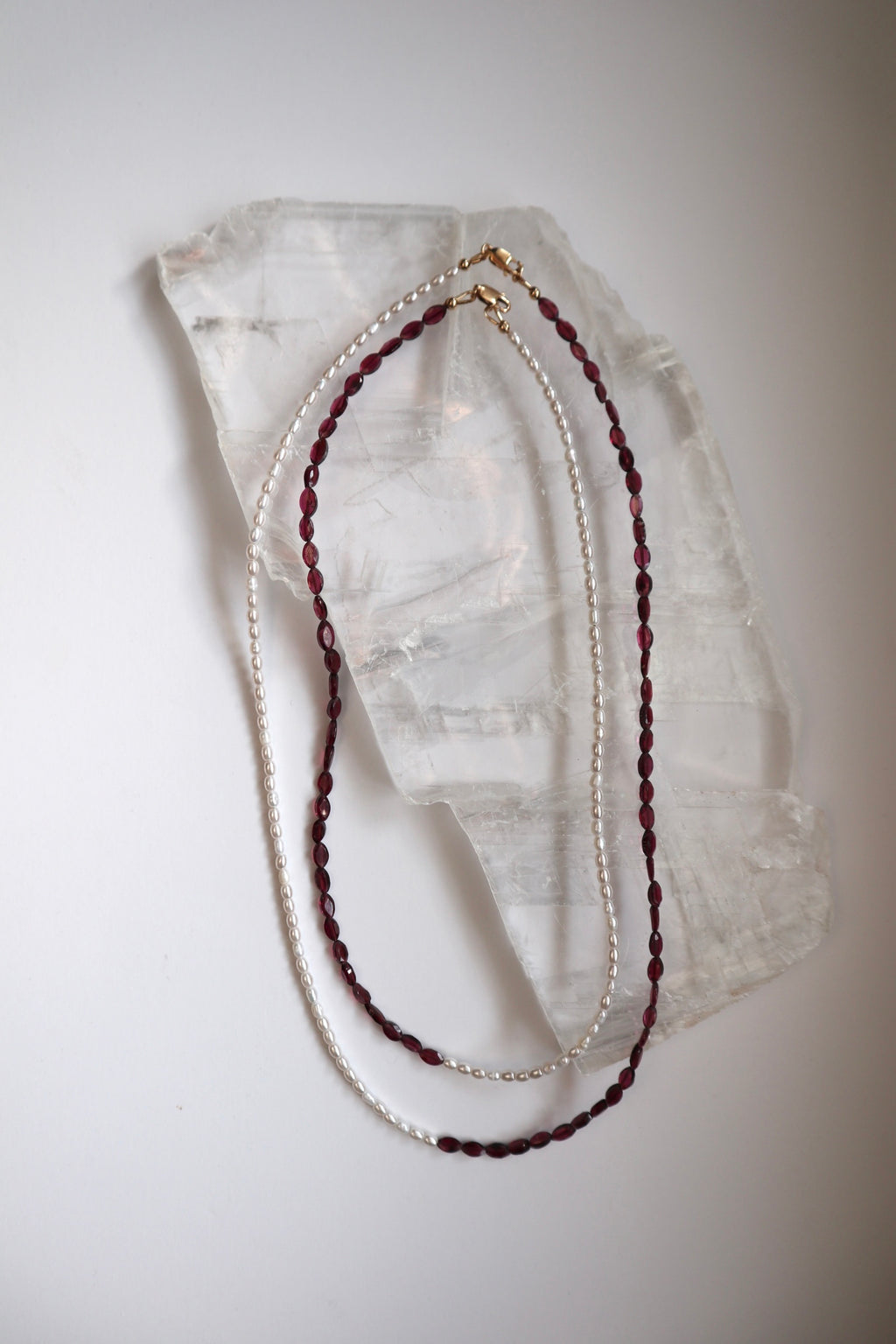 Garnelle necklace by Auréad Designs in both available lengths draped on a white selenite slab, showing the asymmetrical design with deep red marquise garnet beads and freshwater pearl sections side by side