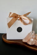 White gift box with gold satin ribbon bow and Auréád Designs logo seal, displayed on a walnut tray alongside a rose quartz crystal cluster.