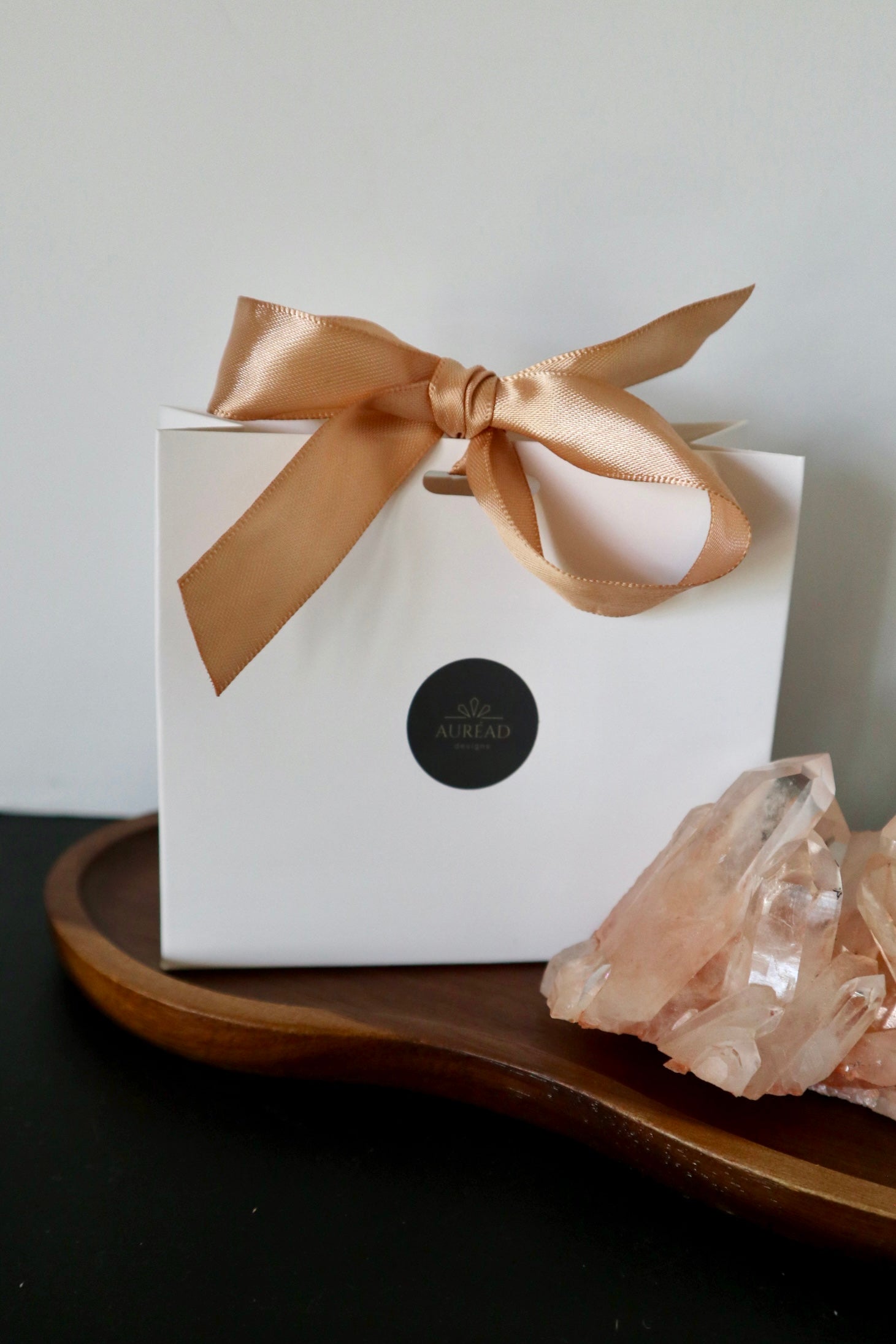 White gift box with gold satin ribbon bow and Auréád Designs logo seal, displayed on a walnut tray alongside a rose quartz crystal cluster.