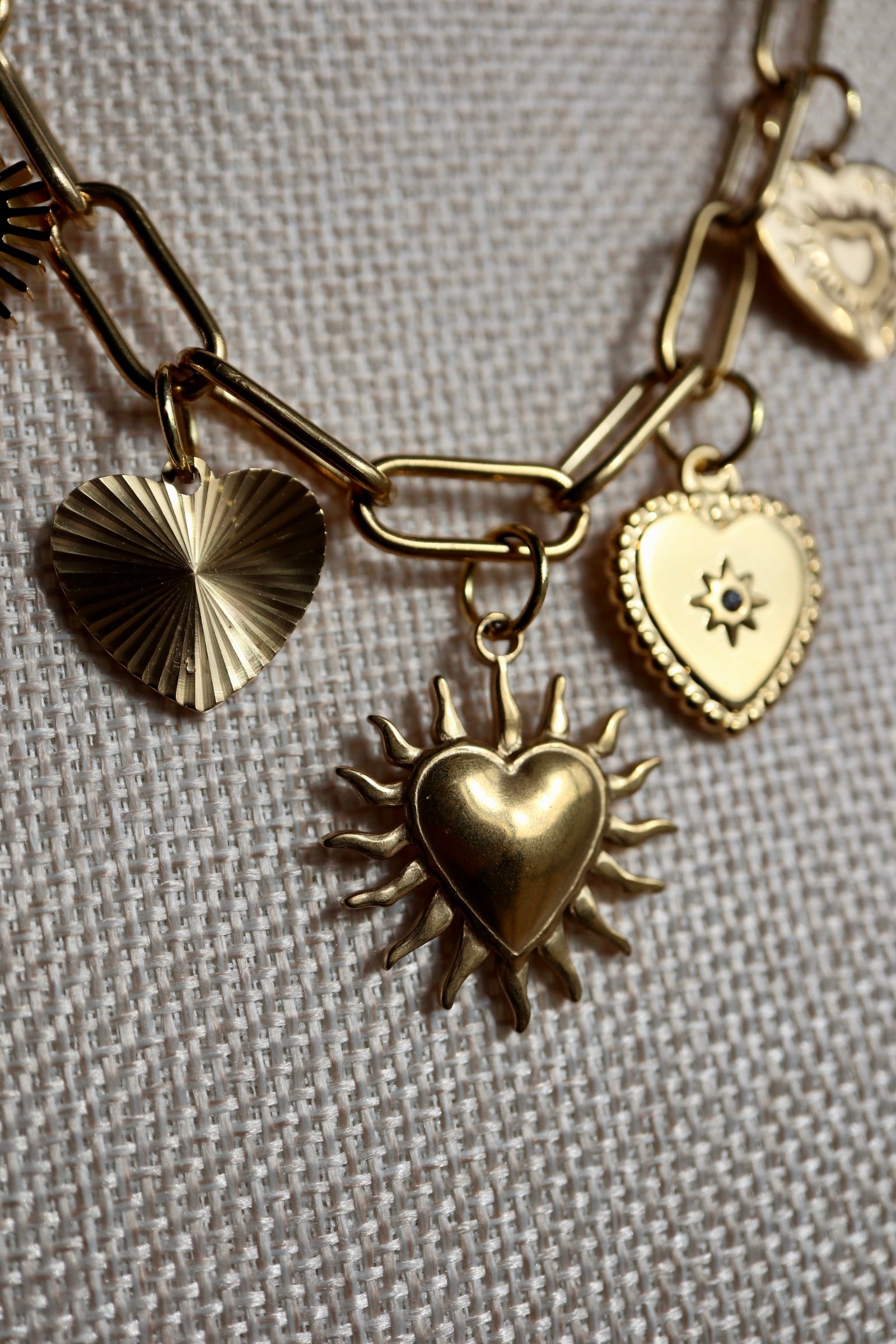 Close-up of the Heartstrong necklace on a linen bust form, showing four gold stainless steel heart charm styles: a ribbed sunburst heart, a sacred heart with rays, a beaded-edge heart with star detail, and a filigree heart, suspended from the gold paperclip chain