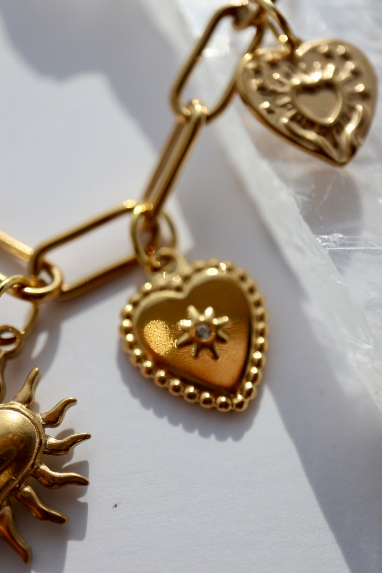 Macro close-up of the Heartstrong necklace by Auréad Designs showing three gold stainless steel charm styles: a beaded-edge heart with sun detail, a raised filigree heart, and a sun charm, suspended from the gold paperclip chain