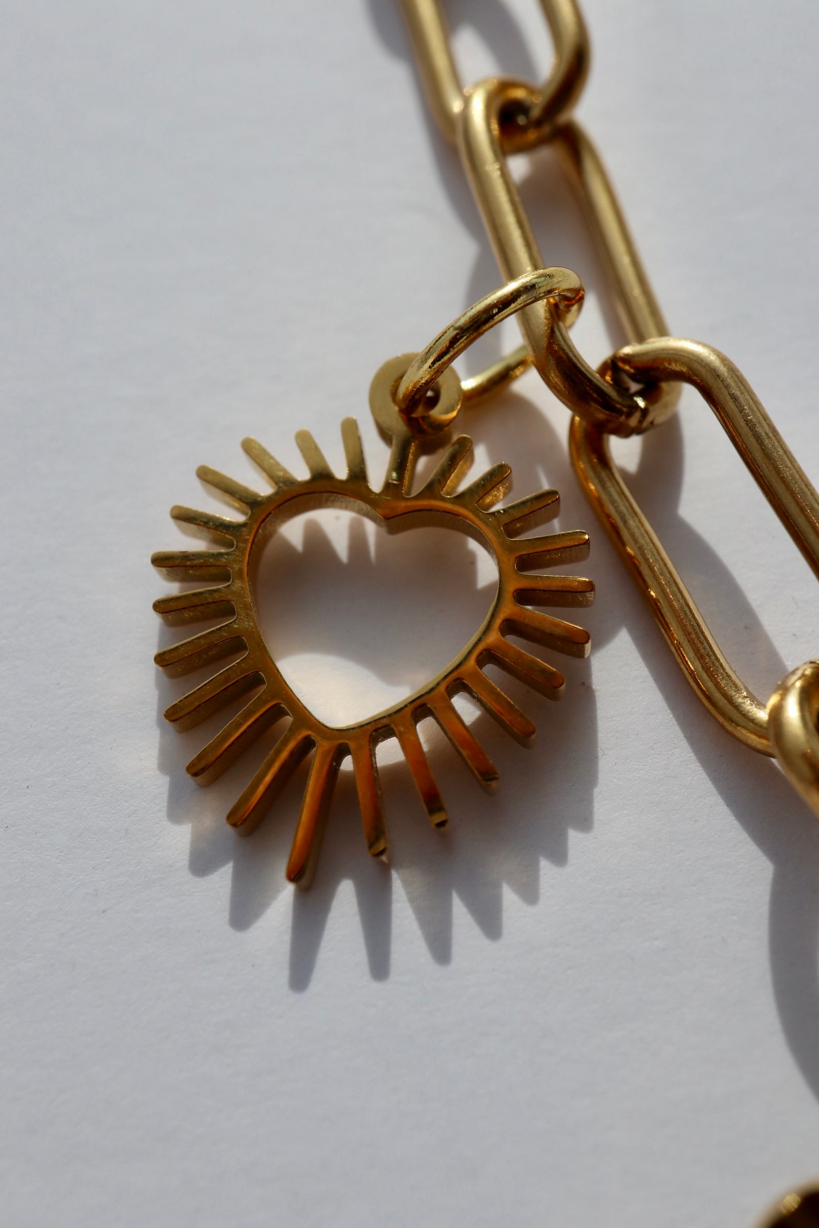 Macro close-up of the Heartstrong necklace by Auréad Designs showing an open sunburst-style gold stainless steel heart charm with radiating rays forming the heart silhouette, suspended from the gold paperclip chain