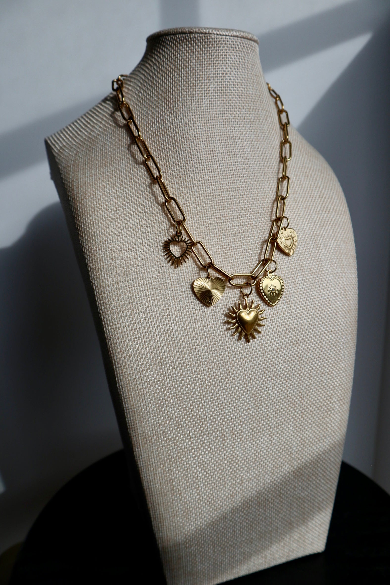 Heartstrong necklace by Auréad Designs displayed on a linen bust form, showing the full gold stainless steel paperclip chain with five varied heart charms in dramatic natural light