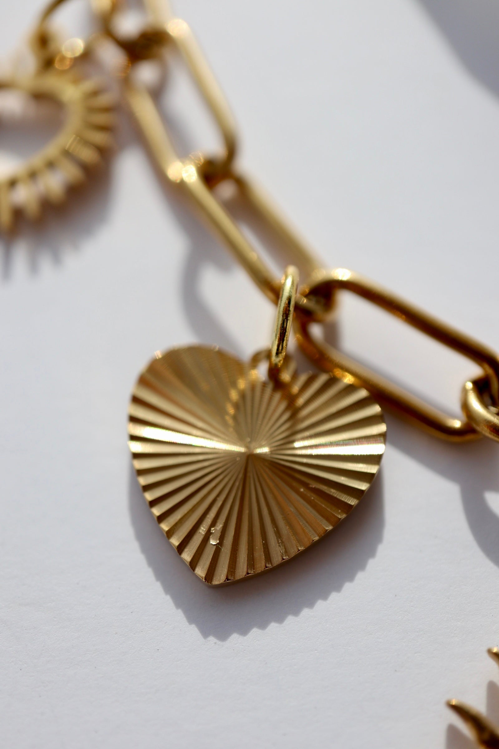 Macro close-up of the Heartstrong necklace by Auréad Designs showing a gold stainless steel ribbed sunburst-pattern heart charm suspended from a gold paperclip chain link