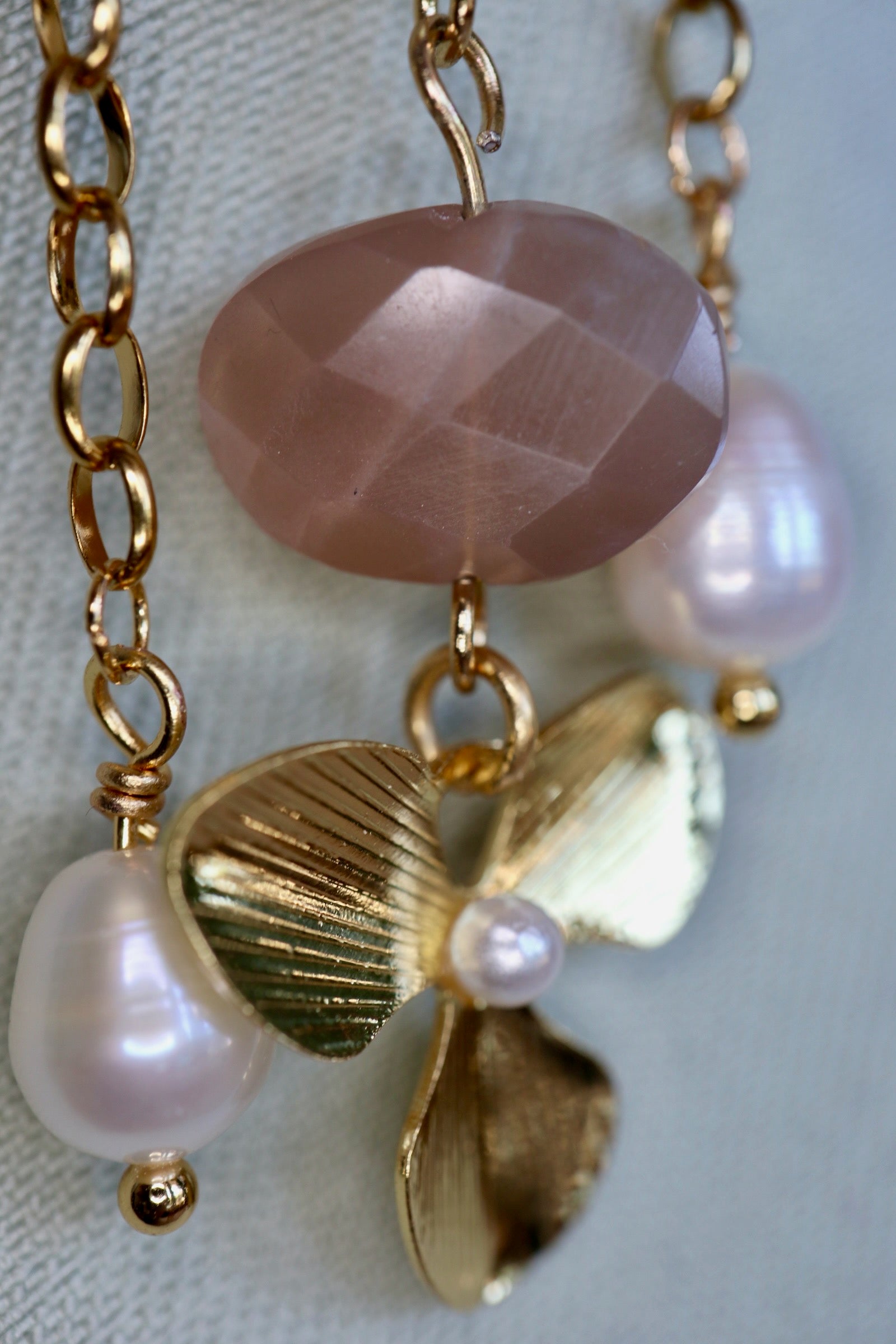 Close-up of Auréad Designs Ilvara brooch showing faceted peach moonstone, freshwater pearl drops, and textured gold three-leaf flower charm on gold cable chain, on a linen bust form.