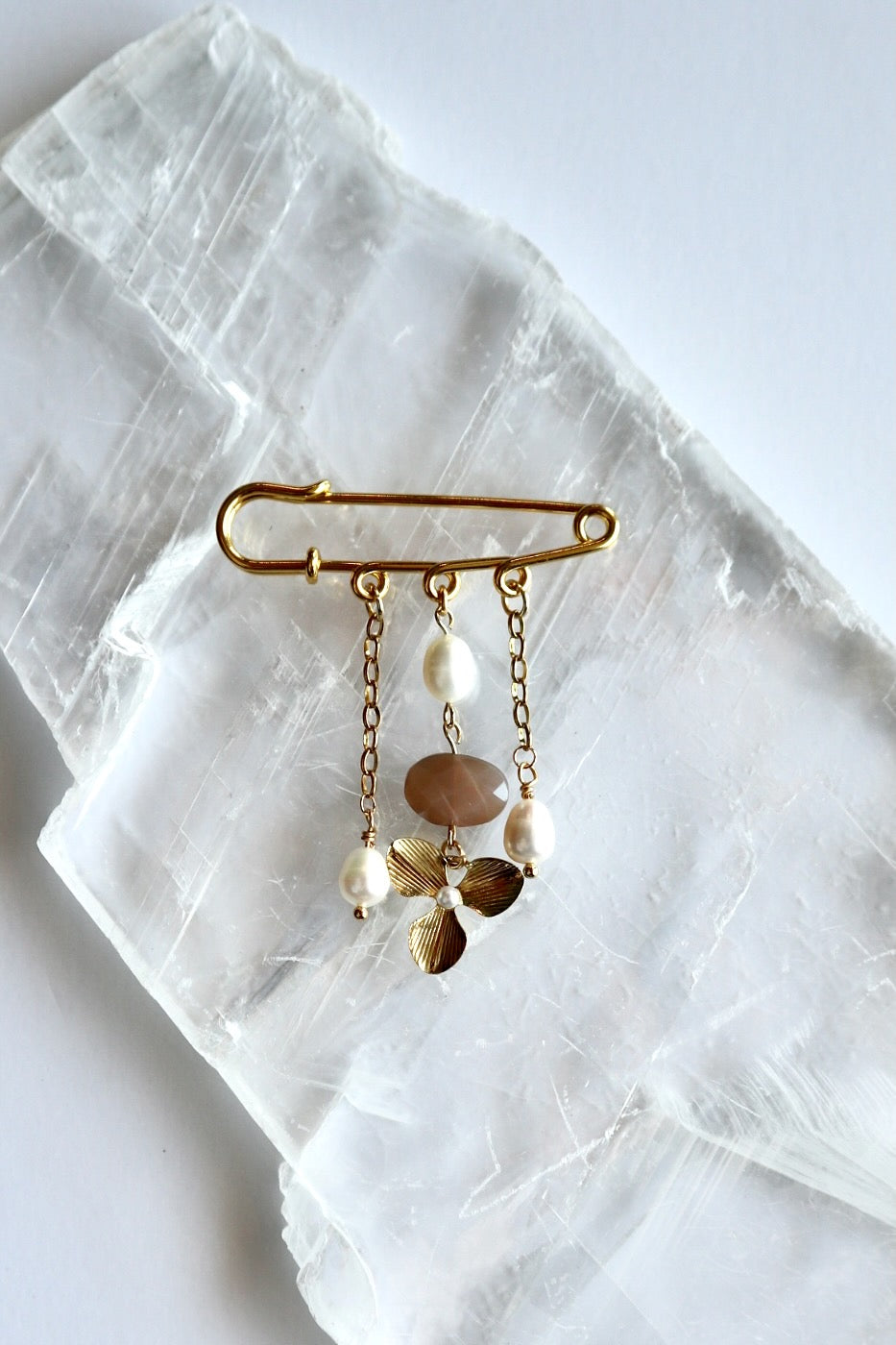 Auréad Designs Ilvara gold kilt pin brooch with peach moonstone, freshwater pearl drops, and gold three-leaf flower charm, flat lay on white selenite slab.