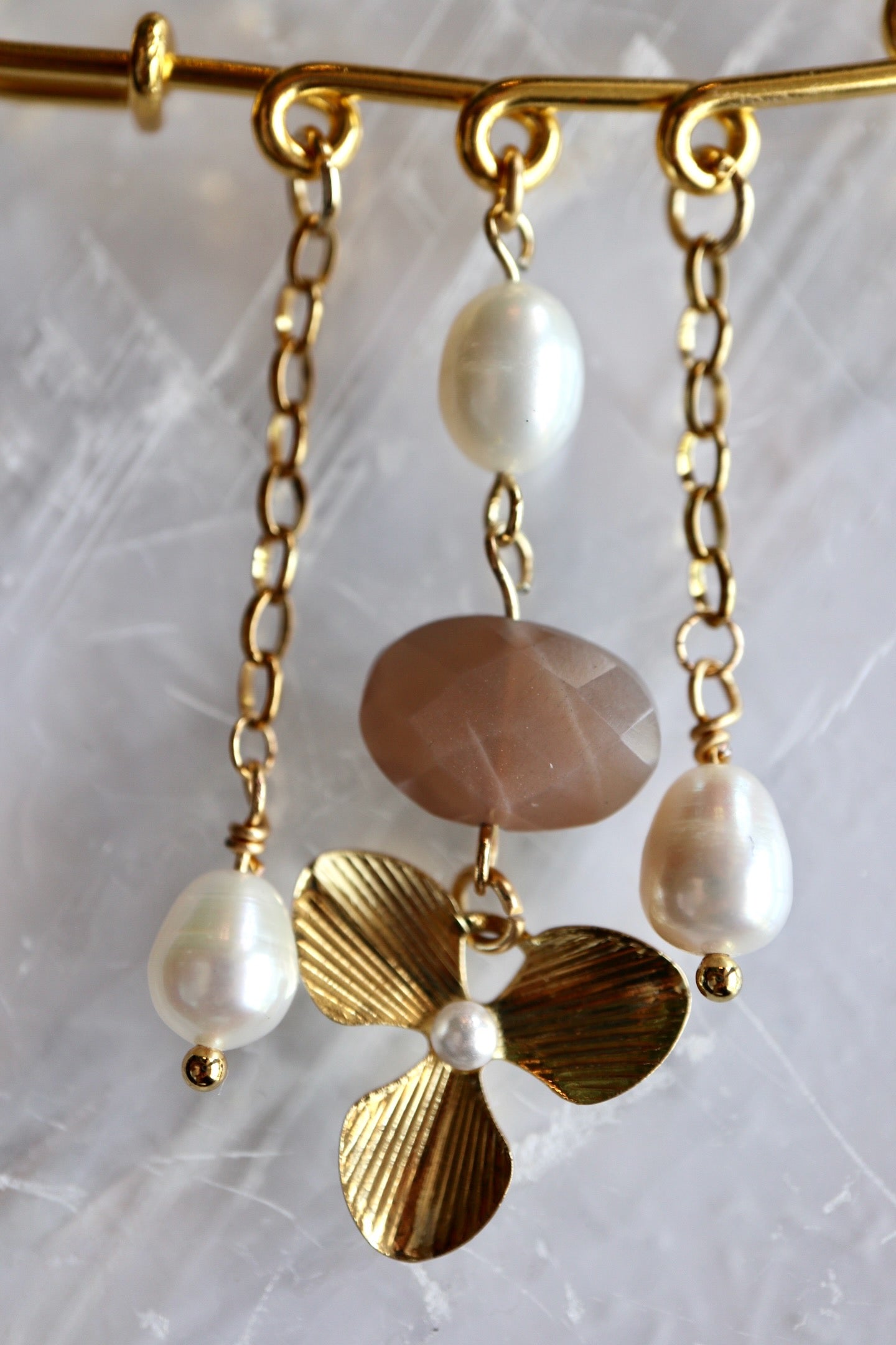 Macro detail of Auréad Designs Ilvara gold kilt pin brooch showing faceted peach moonstone oval, freshwater rice pearls, and gold three-leaf flower charm on gold cable chain drops.