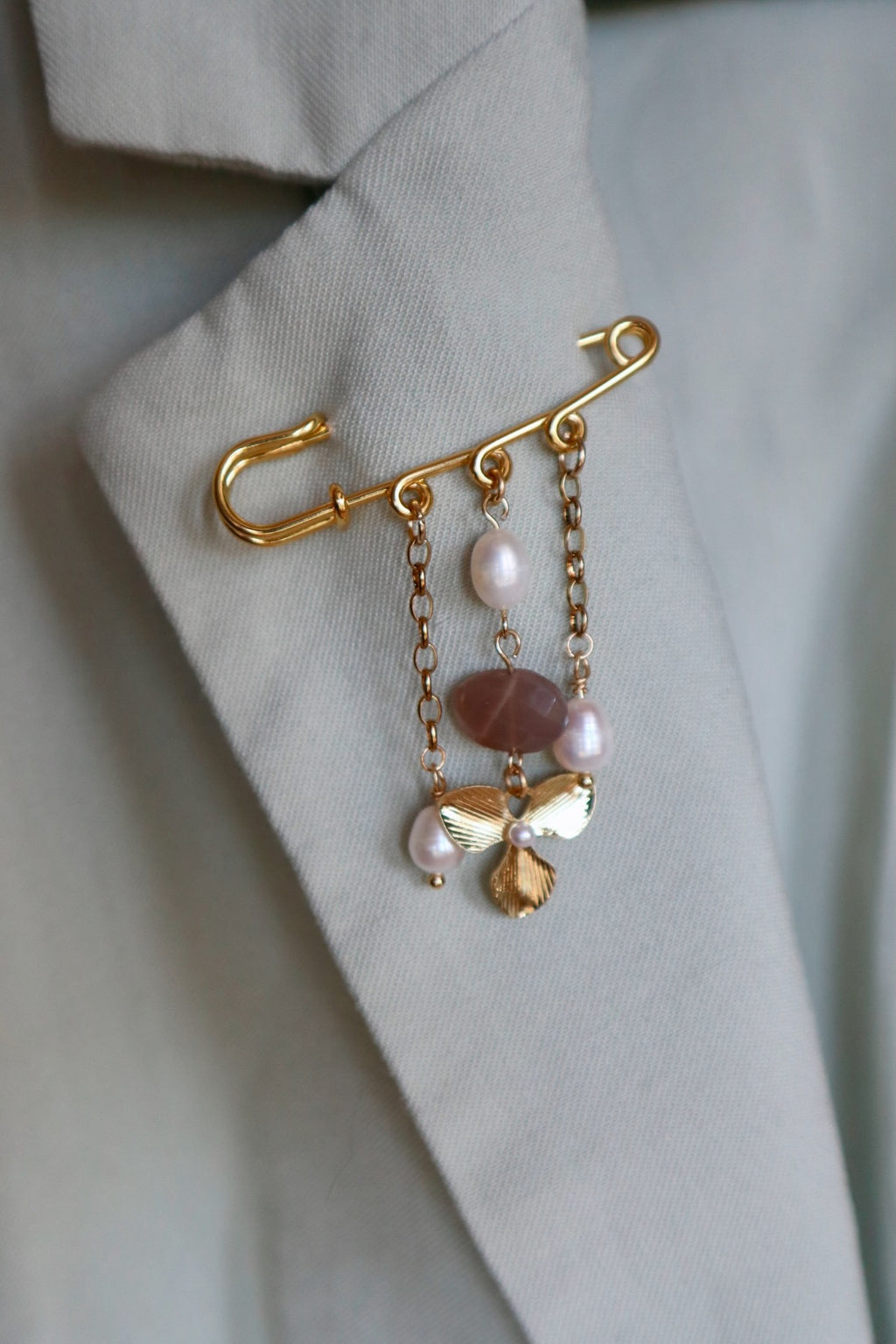 Auréad Designs Ilvara gold kilt pin brooch with peach moonstone and freshwater pearl drops styled on a sage green blazer lapel.