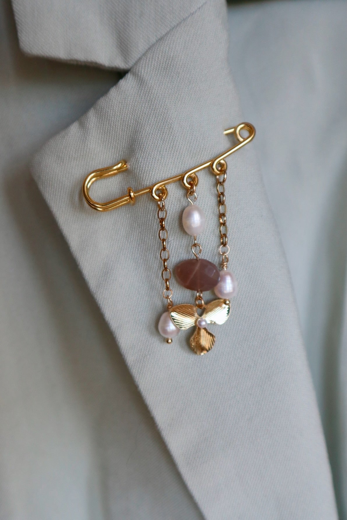 Auréad Designs Ilvara gold kilt pin brooch with peach moonstone and freshwater pearl drops styled on a sage green blazer lapel.