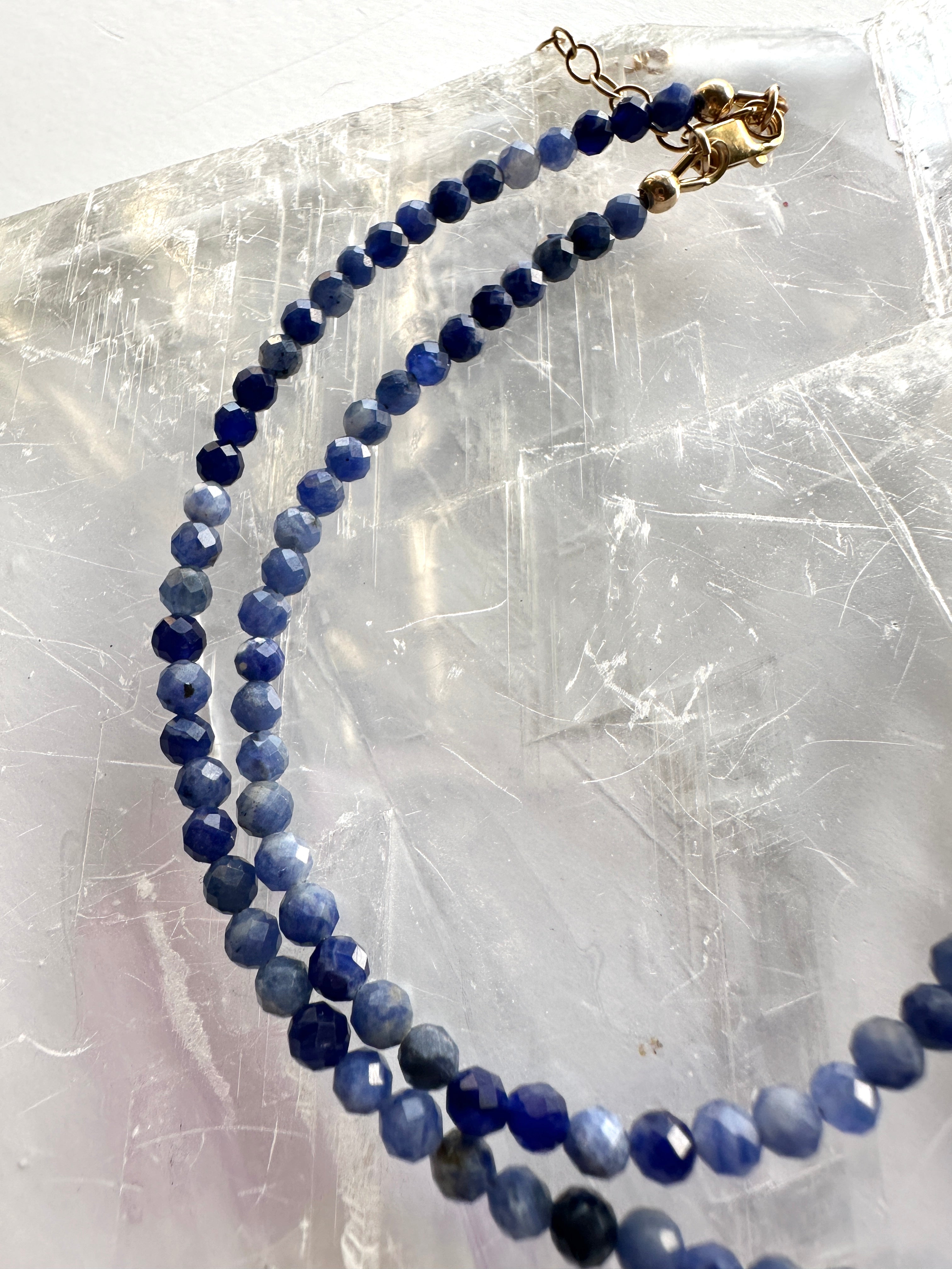 Indira necklace by Auréad Designs in both available lengths coiled on a white selenite slab, showing the natural variation in deep blue sodalite bead tones and the 14k gold-filled lobster clasp and extender chain
