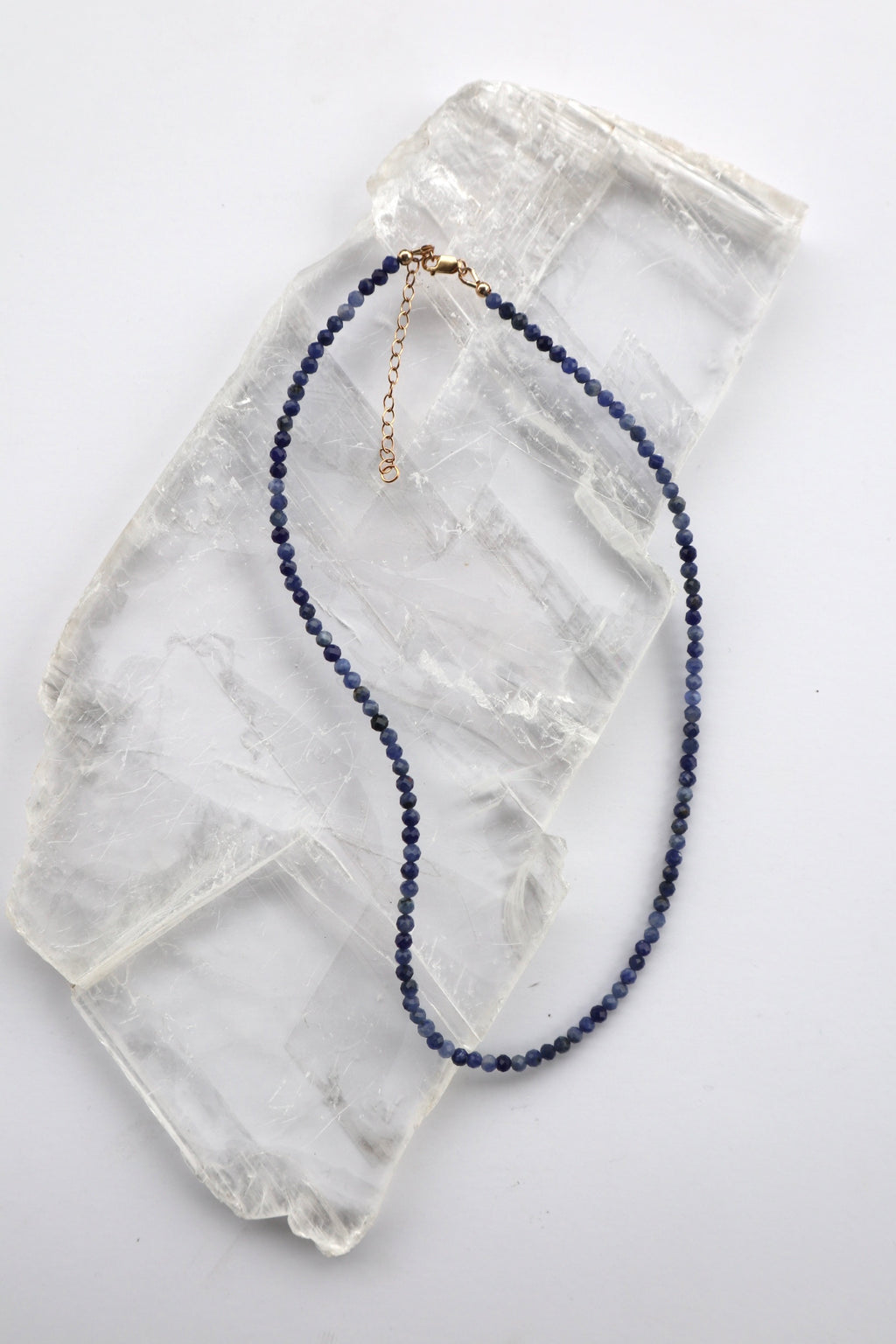  Indira necklace by Auréad Designs, featuring small faceted sodalite gemstone beads in deep blue with a 14k gold-filled clasp and extender chain, laid flat on a white selenite slab