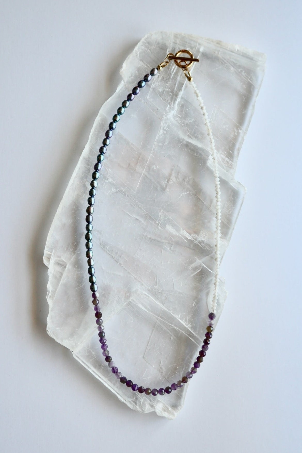 Auréad Designs Irisenne necklace with faceted amethyst beads, peacock green freshwater pearls, and rainbow moonstone rondelles with gold toggle clasp, flat lay on white selenite slab.