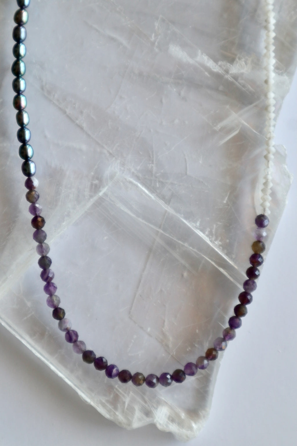Auréad Designs Irisenne necklace showing faceted amethyst and rainbow moonstone bead sections on white selenite slab, with peacock green pearl half visible.