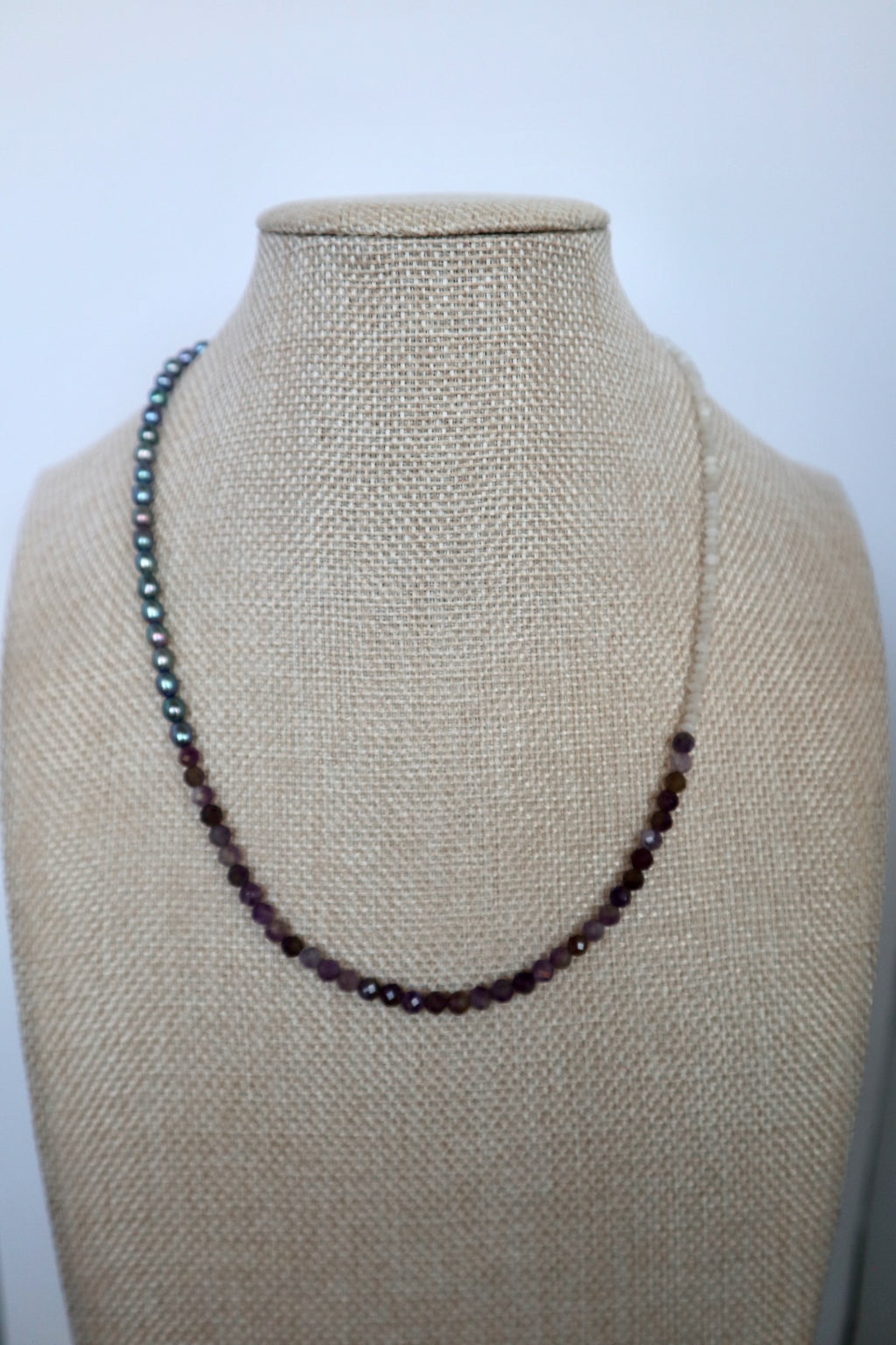 Auréad Designs Irisenne necklace with peacock green freshwater pearls, faceted amethyst, and rainbow moonstone beads displayed on a linen bust form, front view.