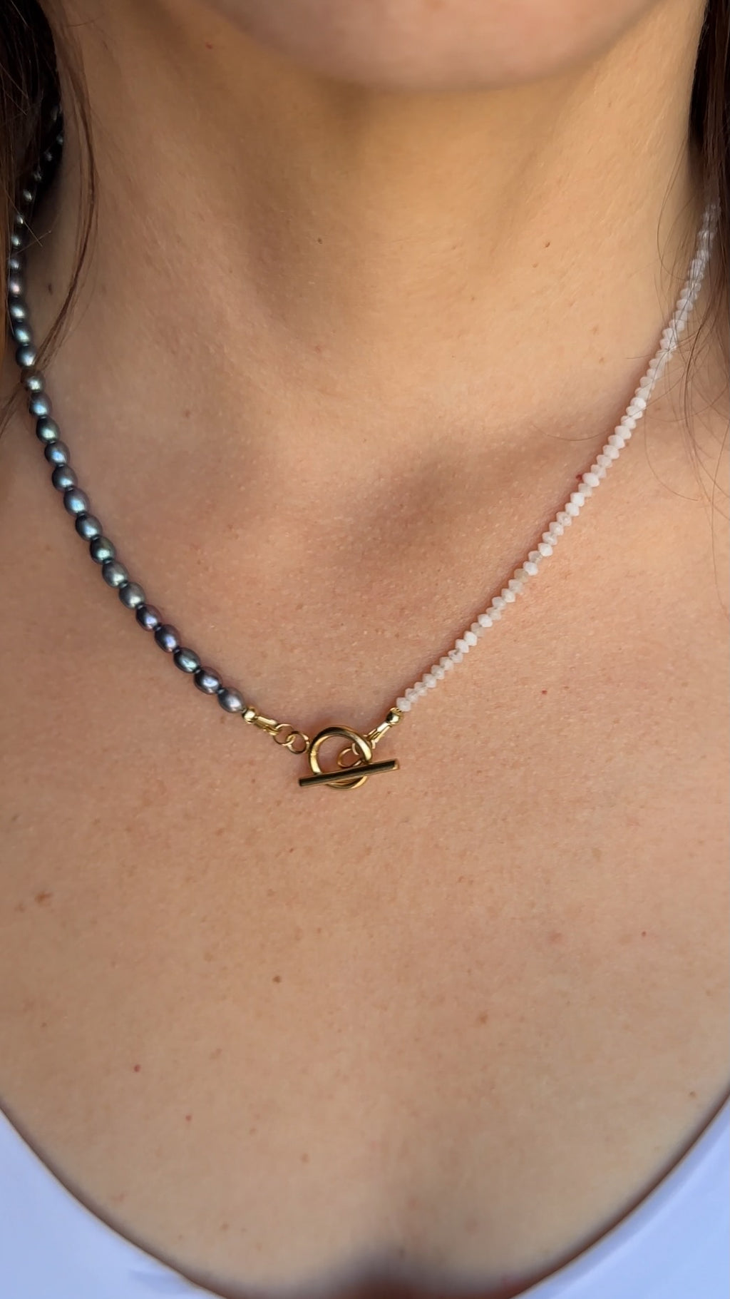 Auréad Designs Irisenne necklace with faceted amethyst beads and peacock green freshwater pearls worn at the collarbone over a white top, gold toggle clasp visible at center front.