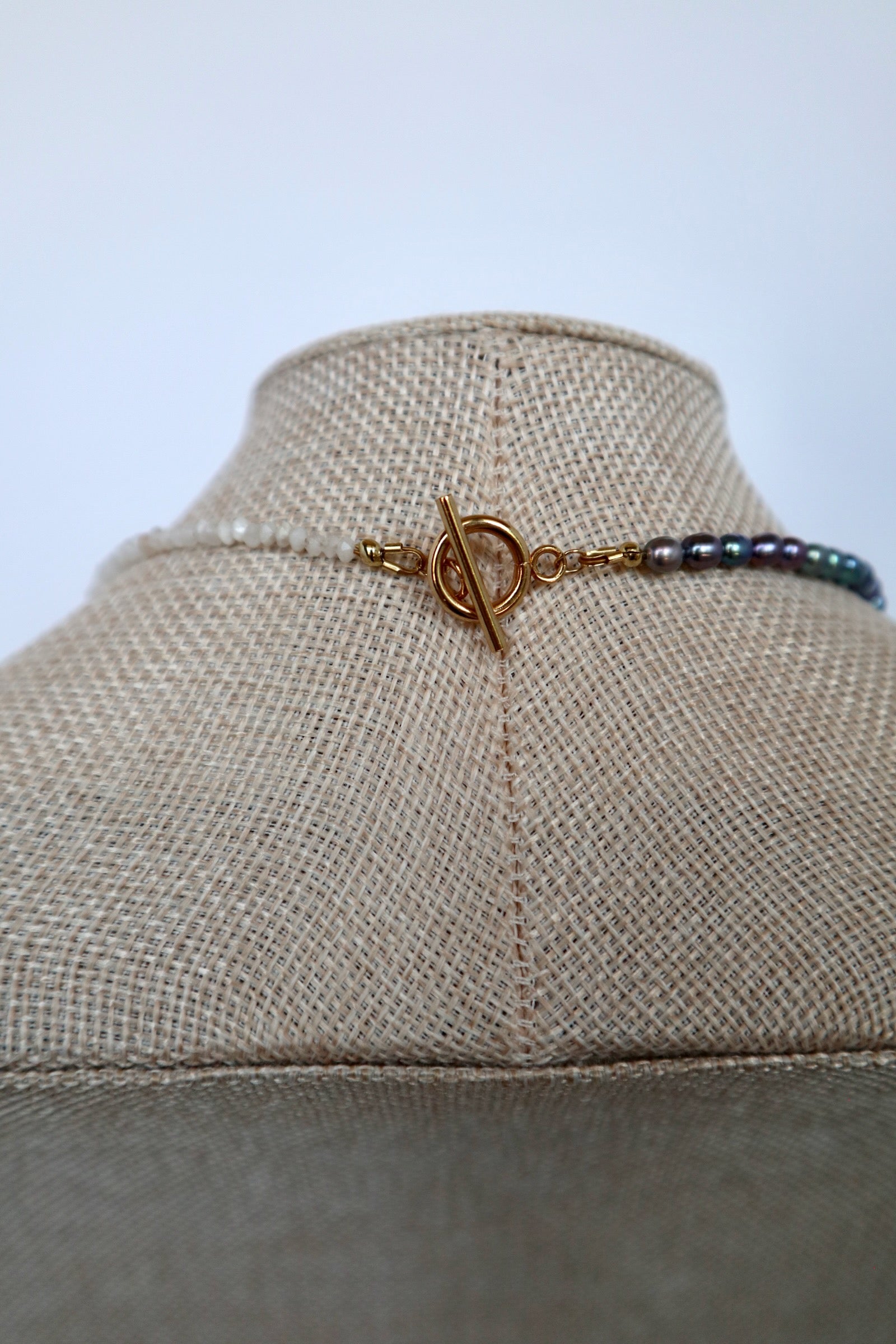 Close-up of Auréad Designs Irisenne necklace gold toggle clasp with peacock green freshwater pearl and rainbow moonstone beads on a linen bust form.
