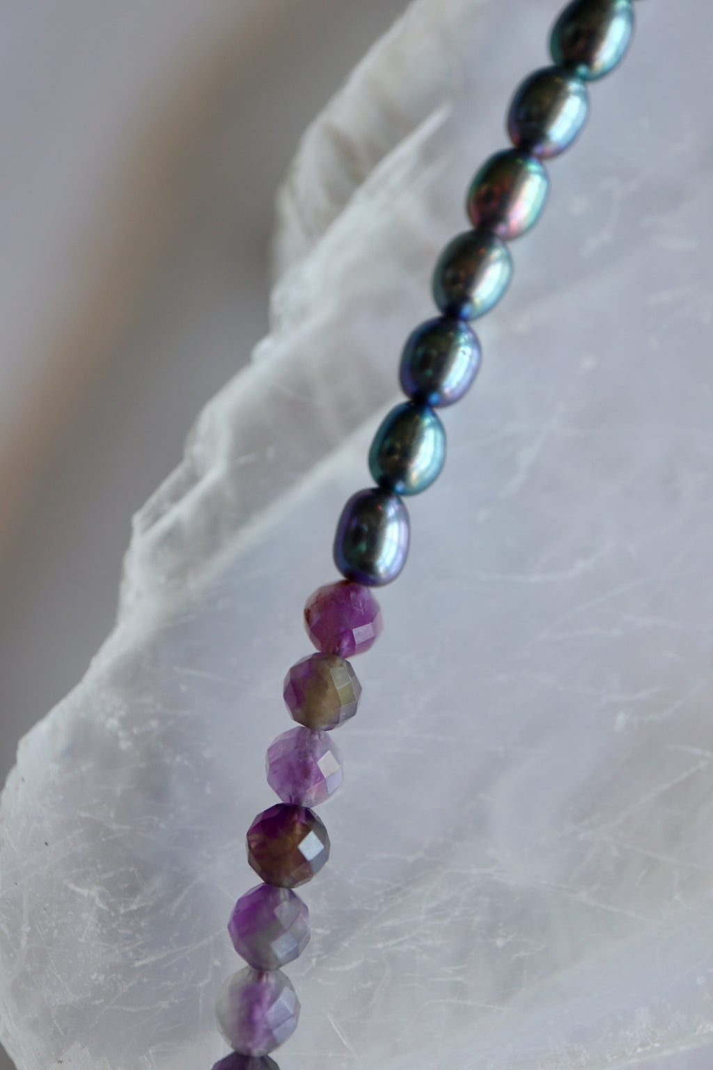Macro detail of Auréad Designs Irisenne necklace showing the transition from iridescent peacock green freshwater pearls to faceted amethyst beads on white selenite slab.
