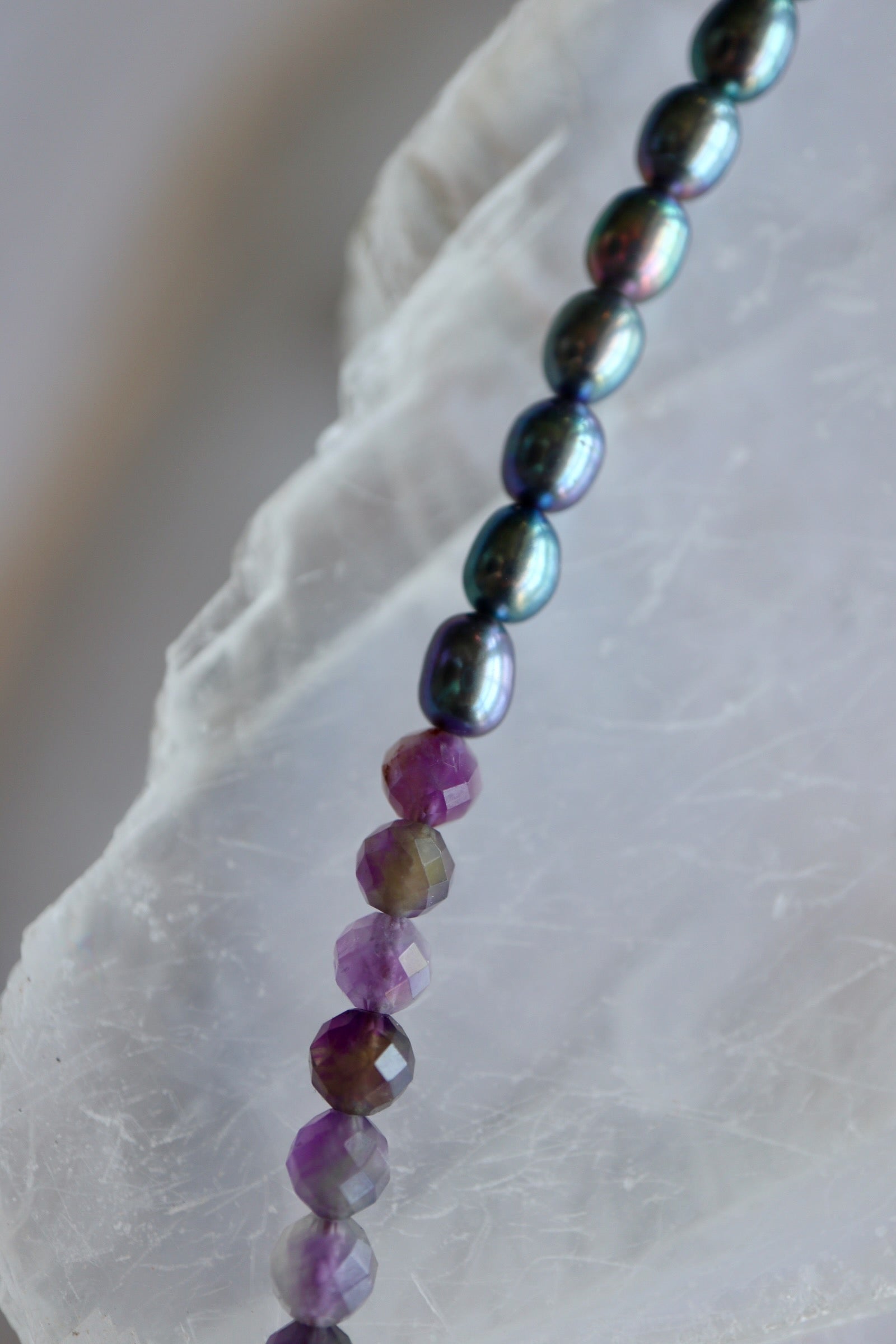 Macro detail of Auréad Designs Irisenne necklace showing the transition from iridescent peacock green freshwater pearls to faceted amethyst beads on white selenite slab.