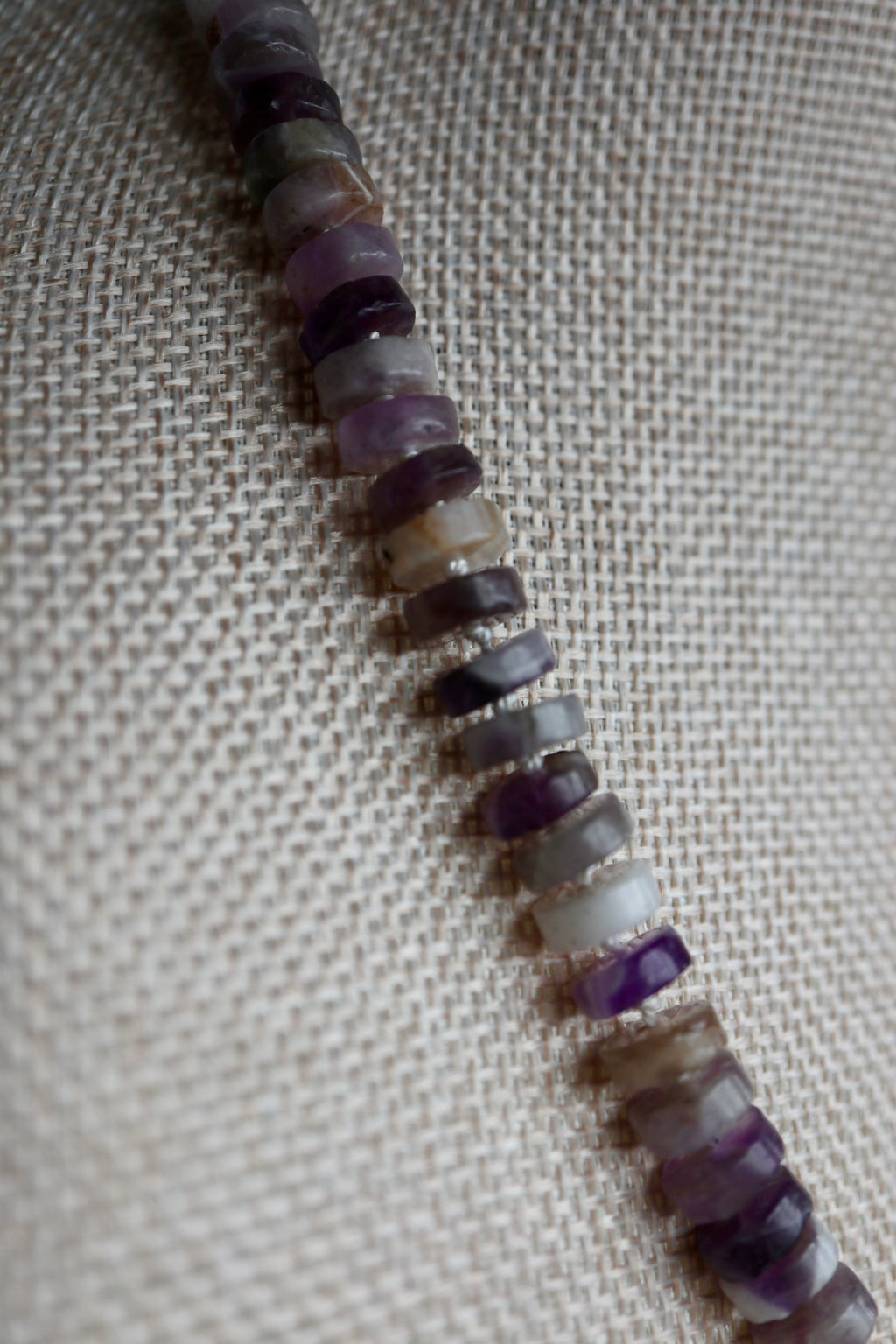 Close-up of the Isolde necklace on a linen bust form, showing the natural color variation of amethyst heishi beads from deep purple to pale lavender against the texture of the linen