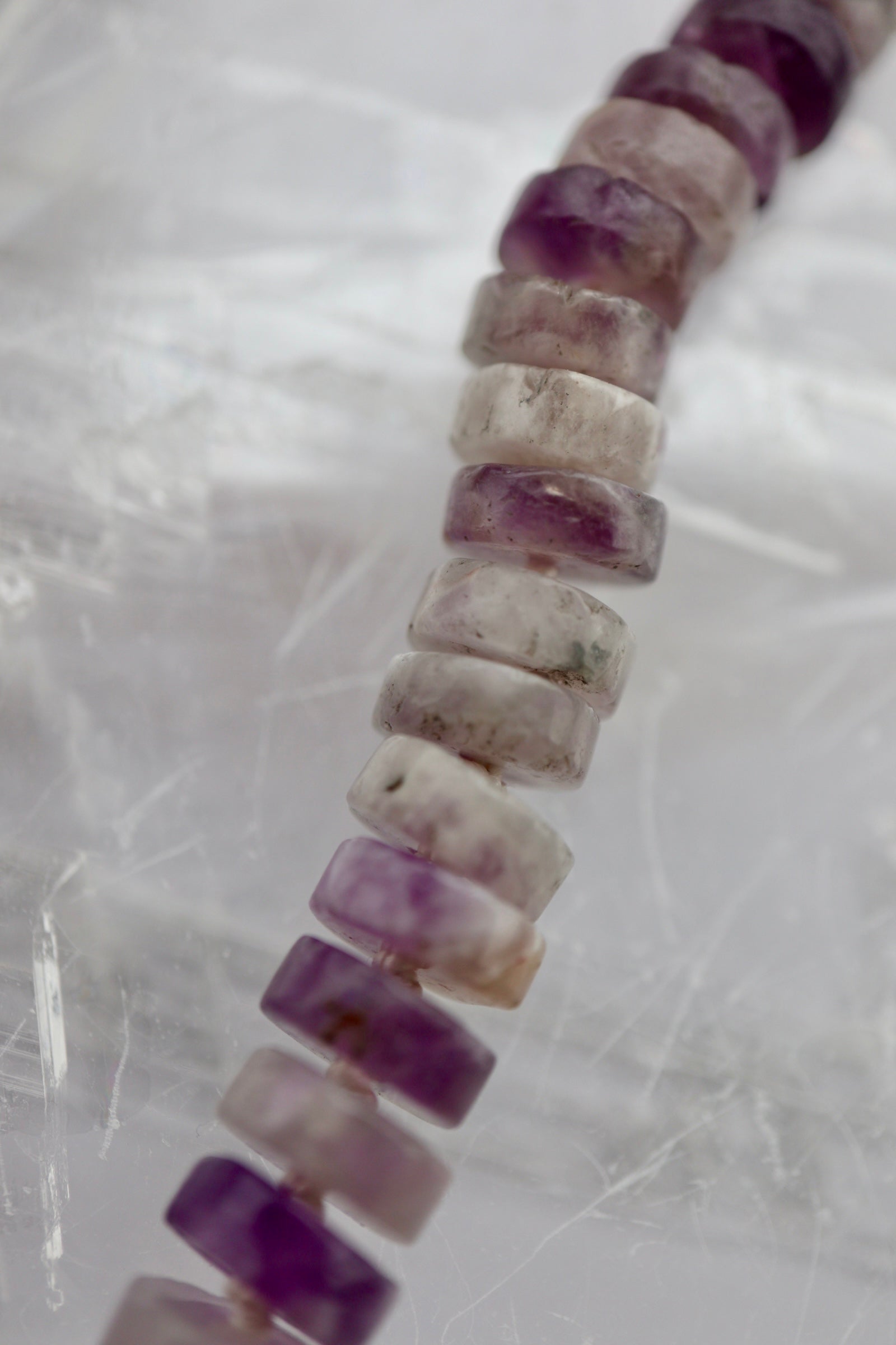 Macro close-up of the Isolde necklace on a white selenite slab, showing the natural color variation of amethyst heishi beads from deep purple to pale lavender and cream, with cream silk hand-knotting between each stone