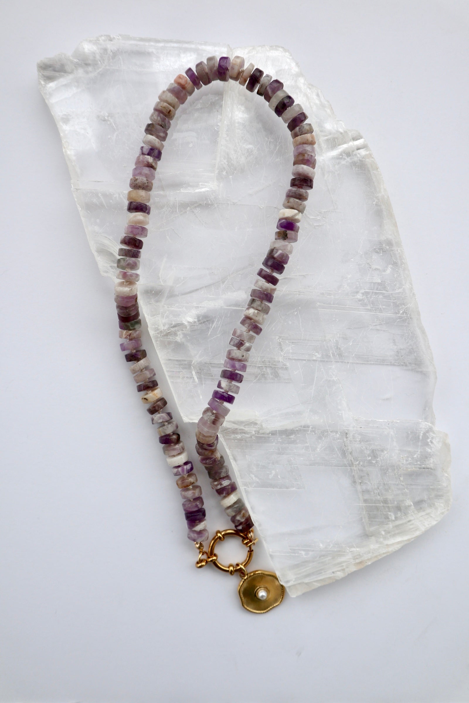 Isolde necklace by Auréad Designs, featuring amethyst heishi beads on a cream silk-knotted strand with a gold stainless steel sailor spring clasp and removable freshwater pearl pendant, laid flat on a white selenite slab