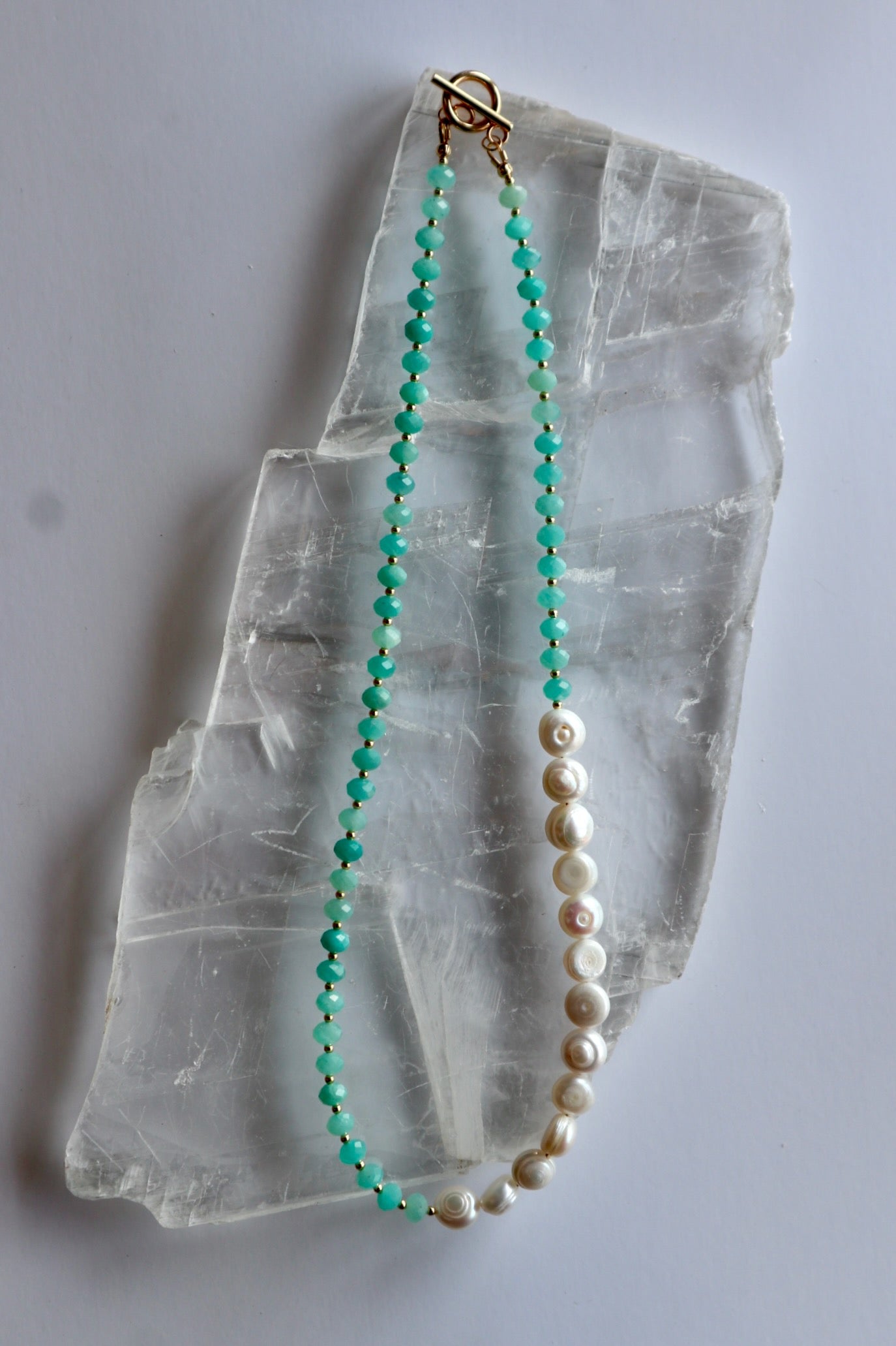 Lake necklace by Auréad Designs laid on a white selenite slab, full view of the asymmetric amazonite rondelle and coin freshwater pearl strand with gold toggle clasp, Glacier Collection