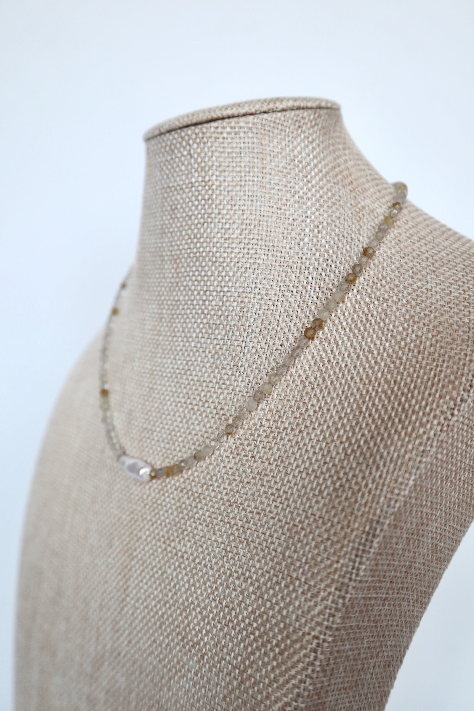 Larch necklace by Auréad Designs on a linen bust, angled view showing the delicate golden rutilated quartz strand and baroque freshwater pearl focal